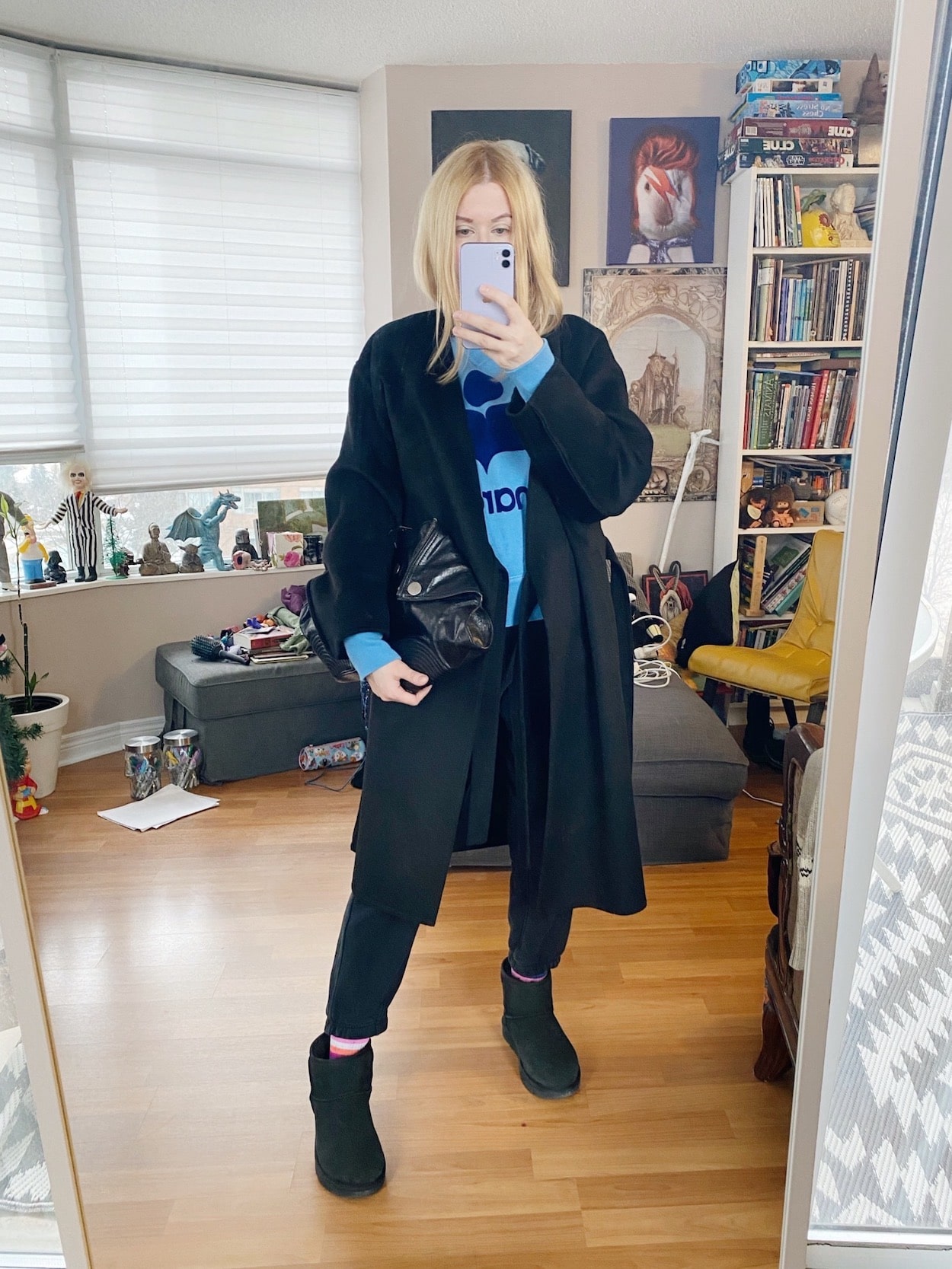 A blonde woman is wearing black joggers, a blue Isabel Marant sweatshirt, a black wrap coat, black Ugg minis, and an Alexander McQueen clutch.