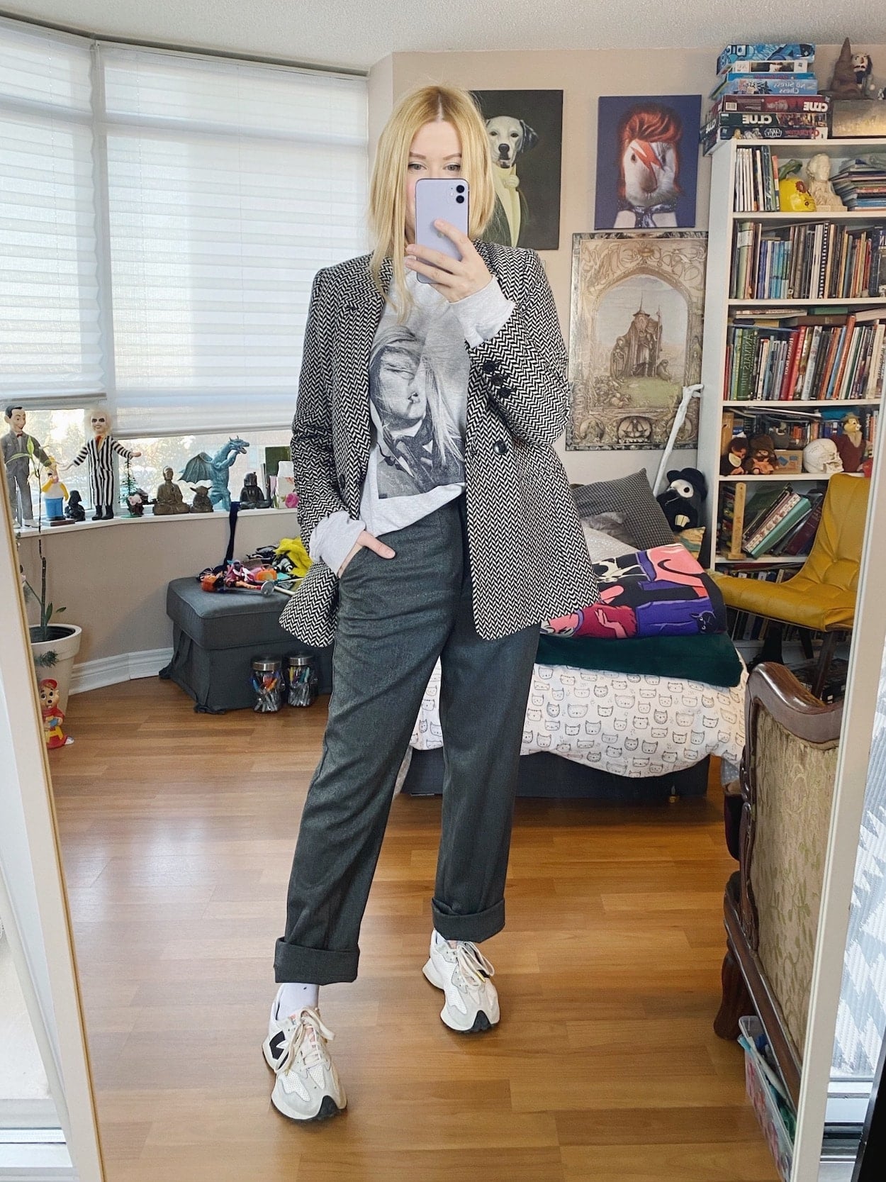 A blonde woman is wearing a grey sweatshirt, grey trousers, a herringbone blazer, and white sneakers.