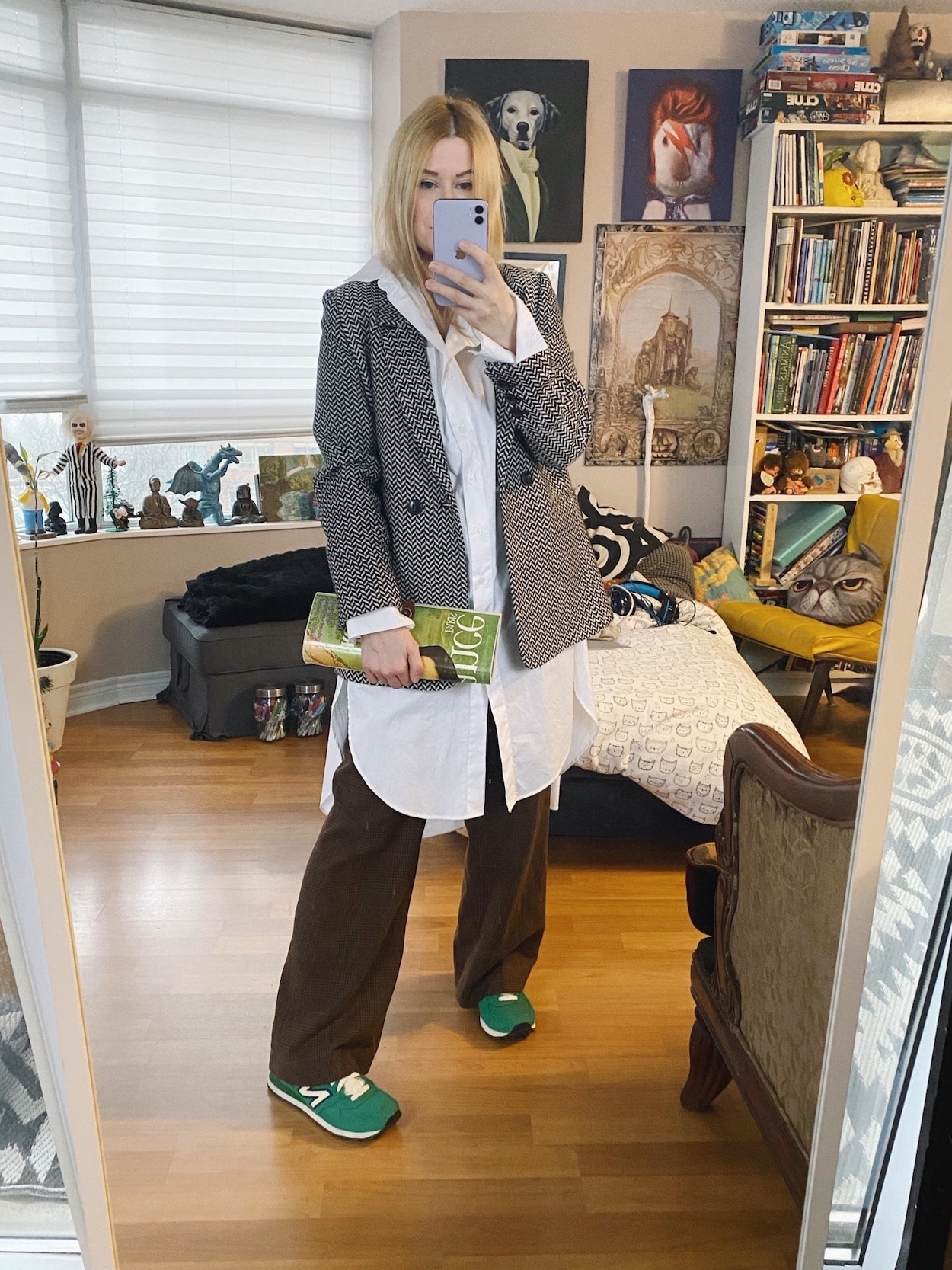 A blonde woman is wearing a long white blouse, over borwn checkered vintage trousers, a herringbone blazer, green sneakers, and a vintage magazine clutch.