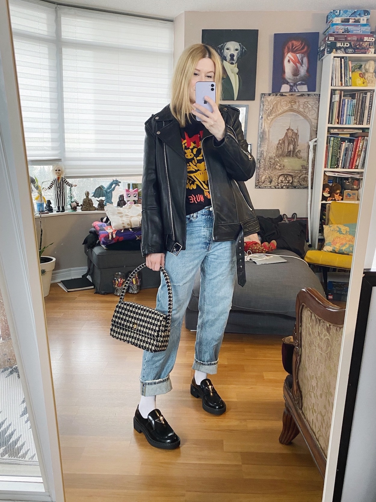 A blonde woman is wearing a Anine Bing sweatshirt, boyfriend jeans, an oversized leather jacket, and a houndstooth handbag.