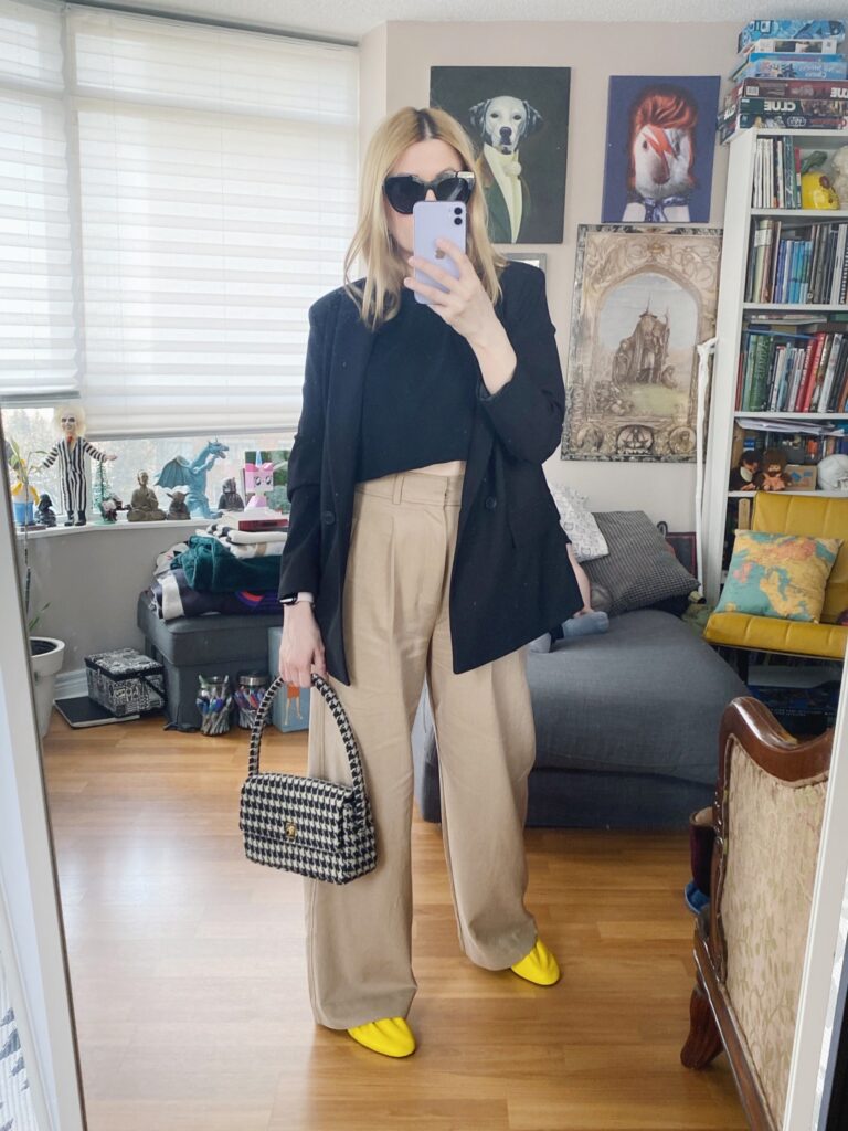 A blonde woman is wearing a black crop t-shirt, tan wide leg trousers, a black blazer, yellow flats, black sunglasses, and a houndstooth purse