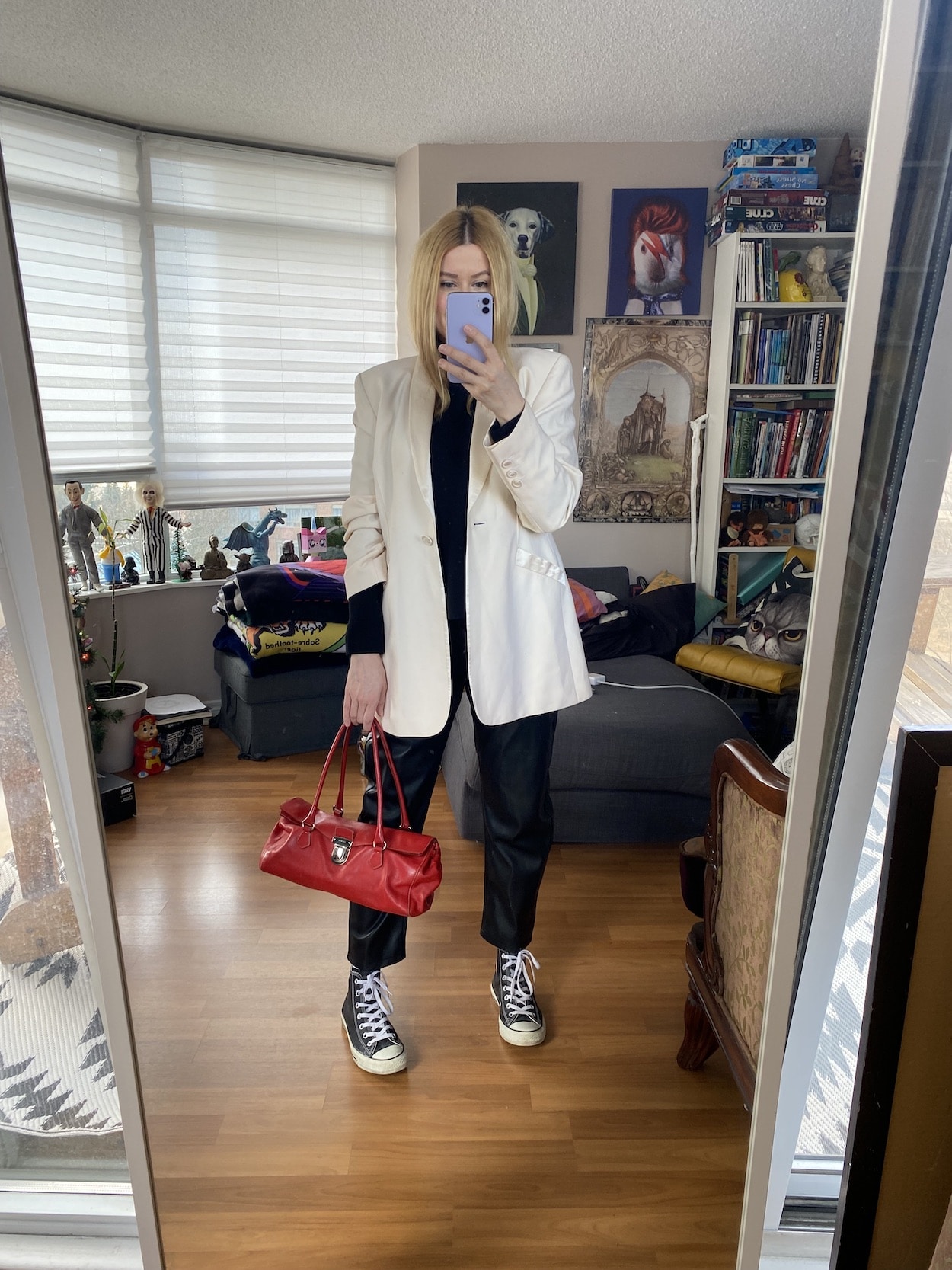 A blonde woman is wearing a black sweater, leather pants, a vintage white blazer, Converse, and a vintage red prada bag.