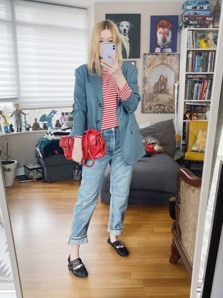 A blonde woman is wearing a red and white striped shirt, a blue-gray blazer, slouchy jeans, Miu Miu flats, and a vintage Prada red purse.