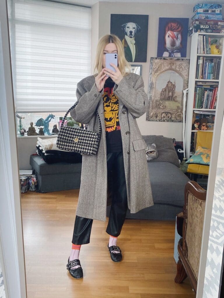 A blonde woman is wearing an Anine BIng sweatshirt, faux leather trousers, a men's herringbone coat, Miu Miu flats, and a handbag.