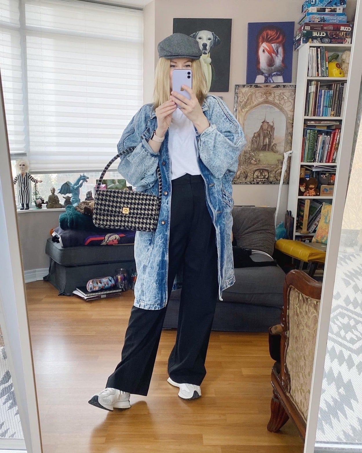 A blonde woman is wearing a white tee. black trousers, a long vintage 80s denim jacket, New Balamce Sneakers, a newsboy cap, and an Isabel Marant purse.