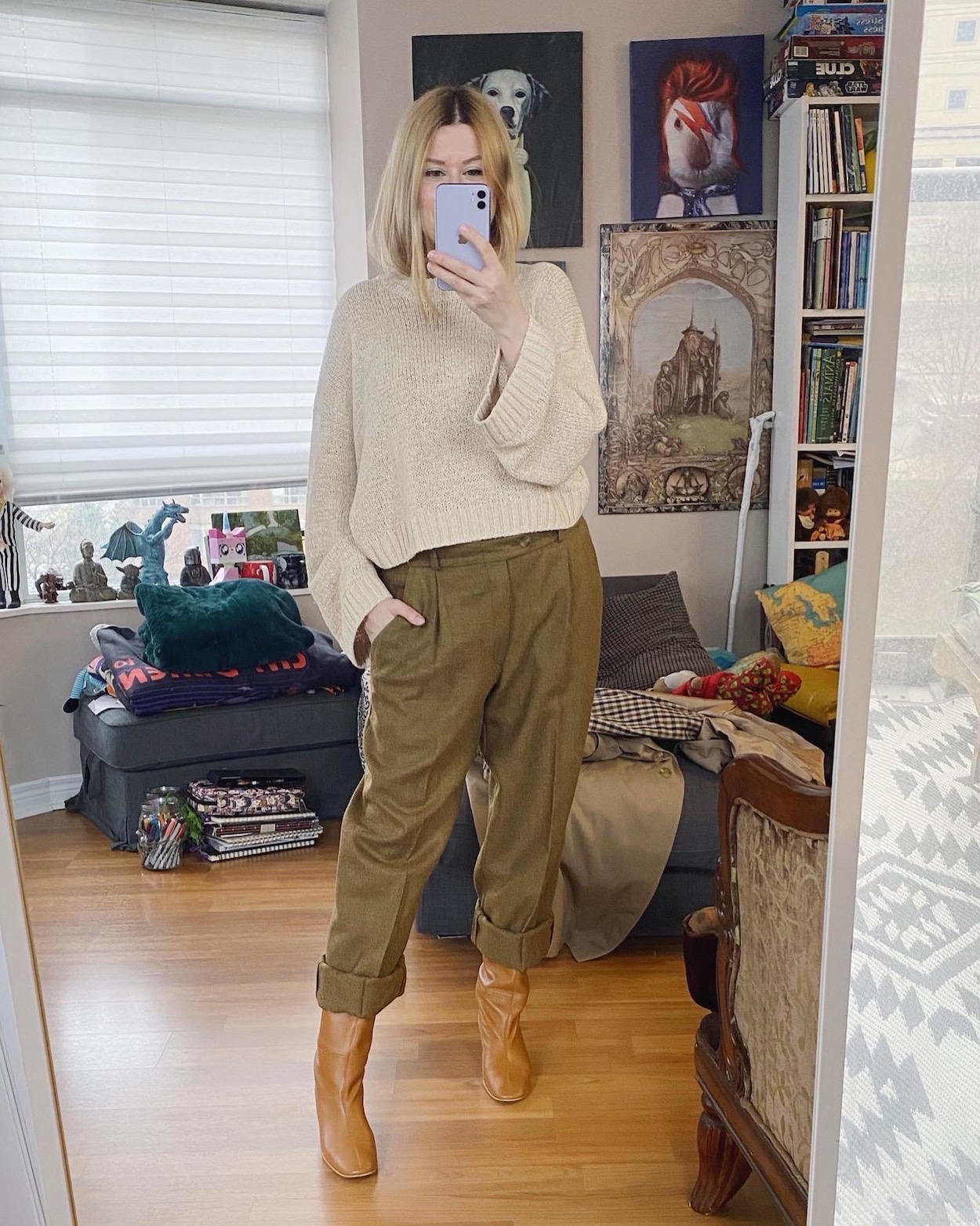 A blonde woman is wearing a cream sweater, vintage trousers, and knee high boots.