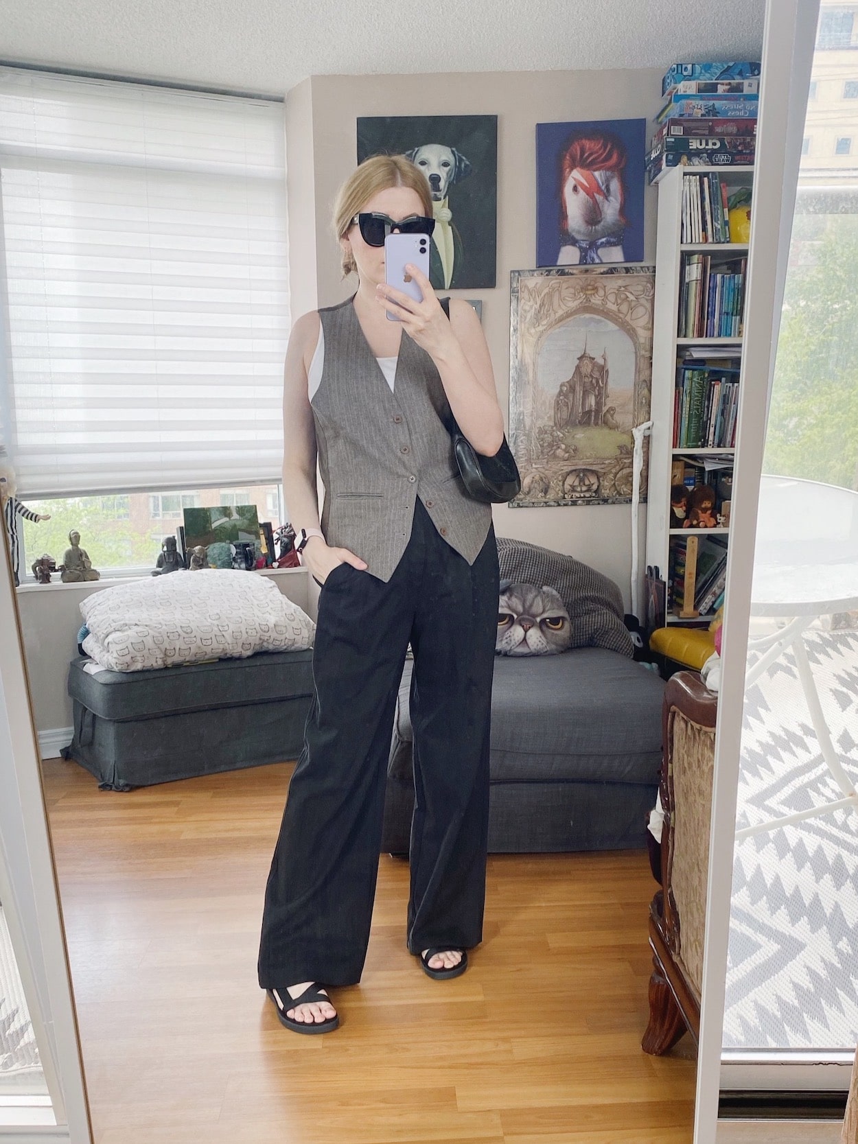 A blonde woman is wearing a vest over a white tank top, black trousers, sports sandlas, black sunglasses, and a vintage Gucci Jackie bag.