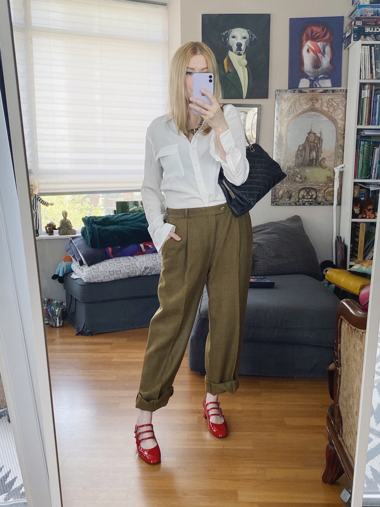 A blonde woman is wearing a white silk blouse, men's vintage trousers, red Carel Kina Mary Janes, a vintage necklace, and a chanel bag.