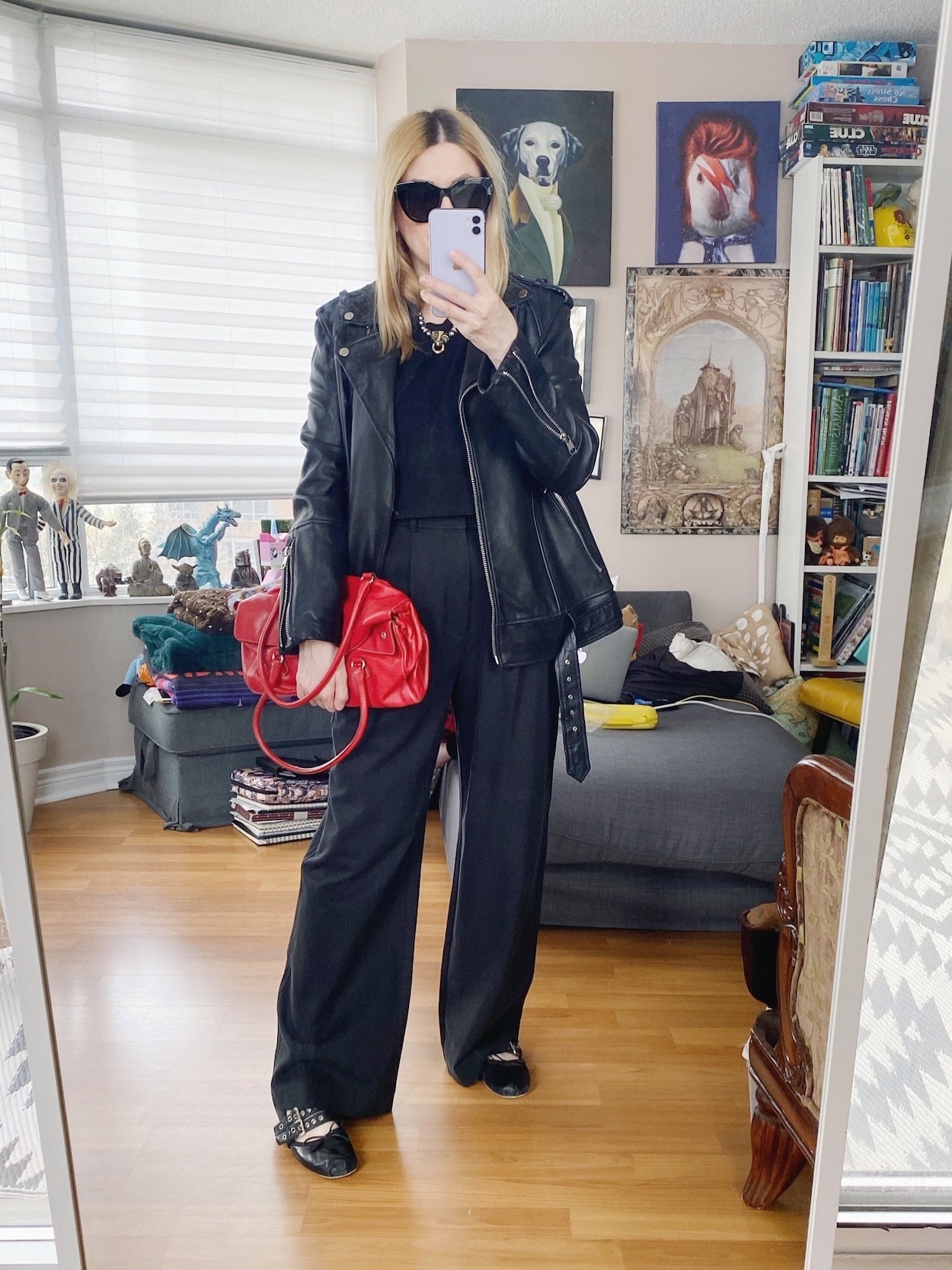 A blonde woman is wearing black trousers, a black crop top, an oversized leather jacket, black sunglasses, black flats, and a red purse.