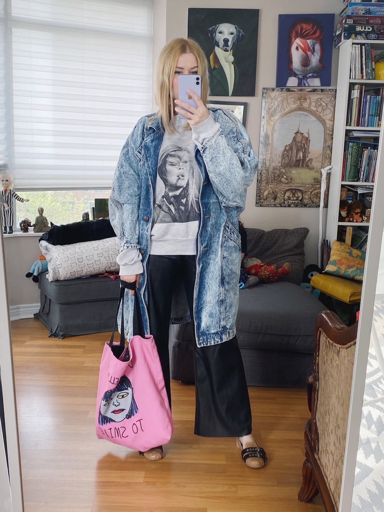 A blonde woman is wearing an Anine Bing sweatshirt, leather trousers, a vintage denim jacket, Miu Miu flats, and a tote.