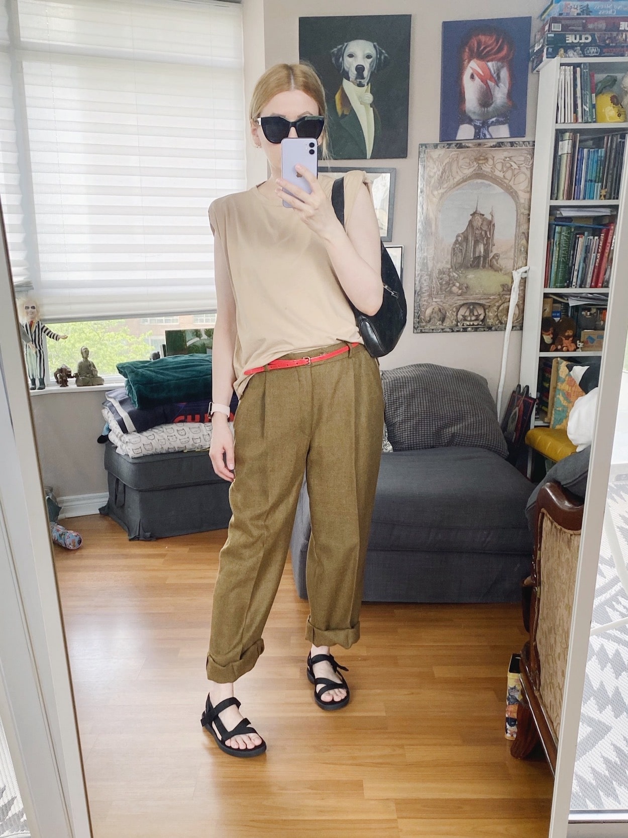 A blonde woman is wearing a shoulder pad tank, vintage trousers, sports sandals, a red belt, black sunglasses, and a vintage Gucci bag.
