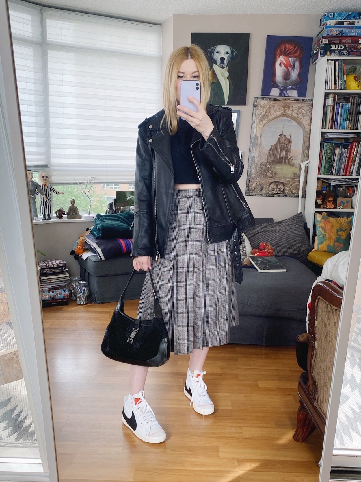 A blonde woman is wearing a black cropped t-shirt, a vintage plaid skirt, and oversized leather jacket, Nike Blazers, and a vintage Gucci bag.