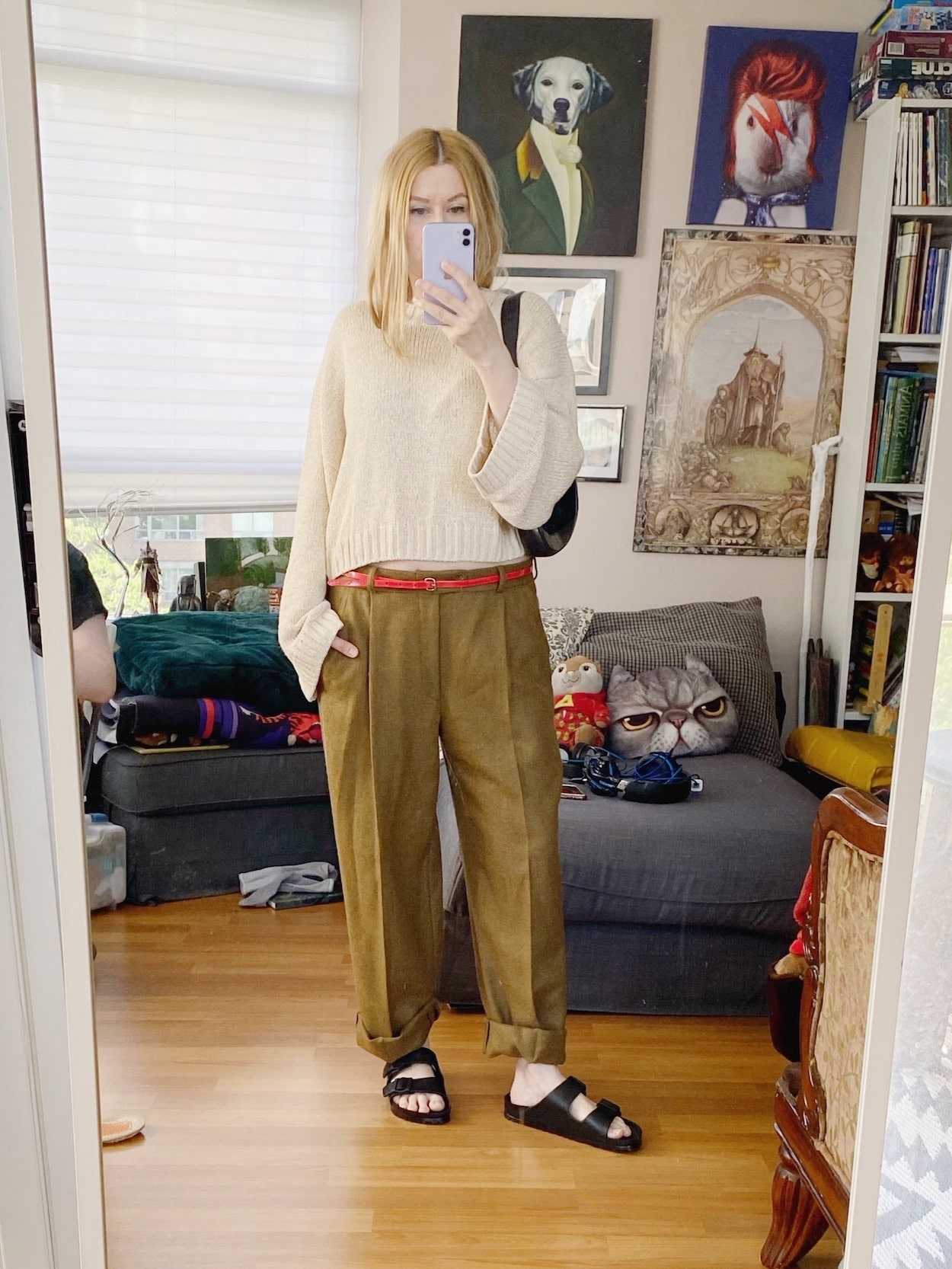 A blonde woman is wearing men's vintage trousers, a red belt, a cropped sweater, Birkenstock sandals, and a vintage Gucci bag.