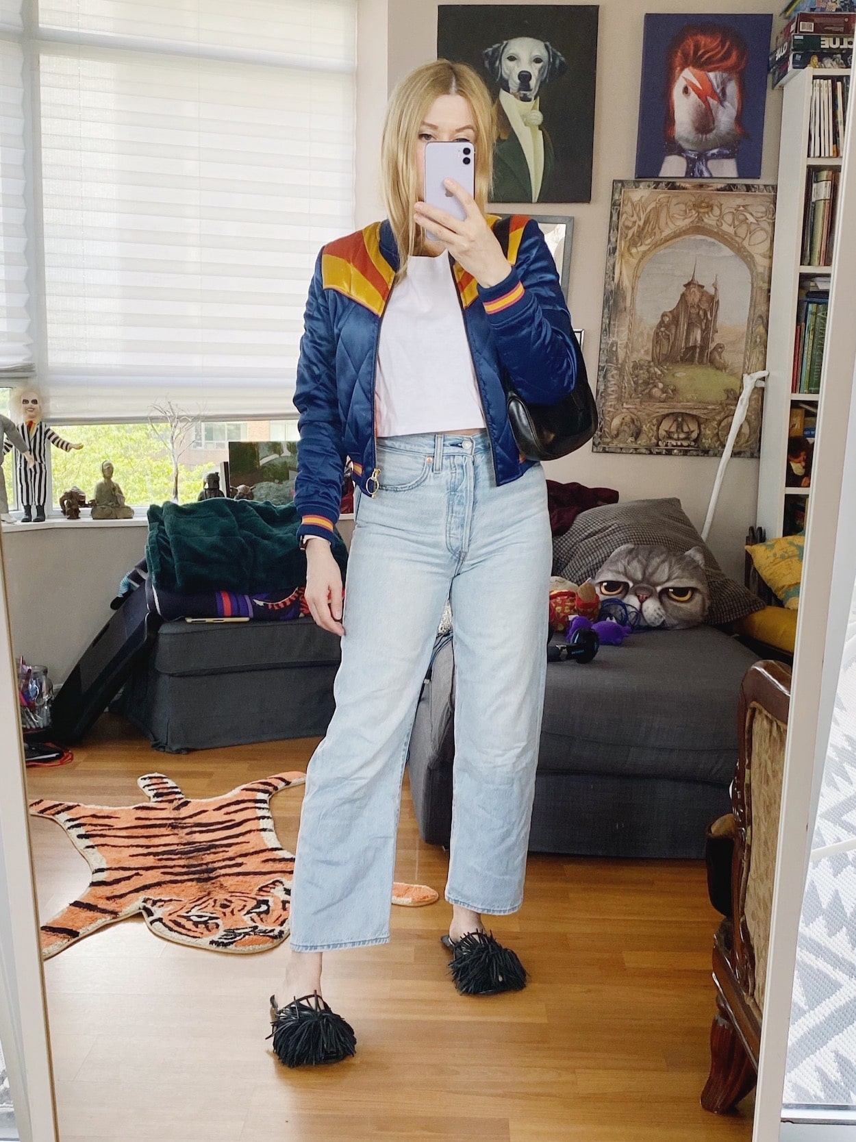 A blonde woman is wearing a retro bomber jacket, white cropped top, Levis, slides, and a vintage Gucci bag.