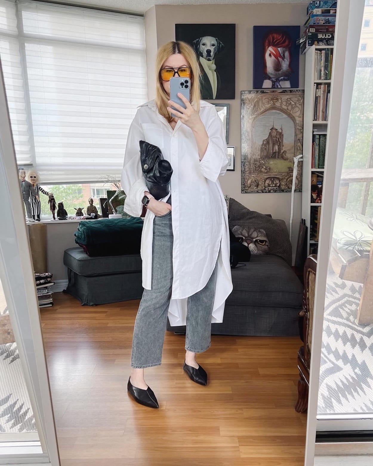 A blonde woman is wearing an oversized button up, grey Agolde jeans, black Acne Studios flats, retro sunglasses, and a black clutch.