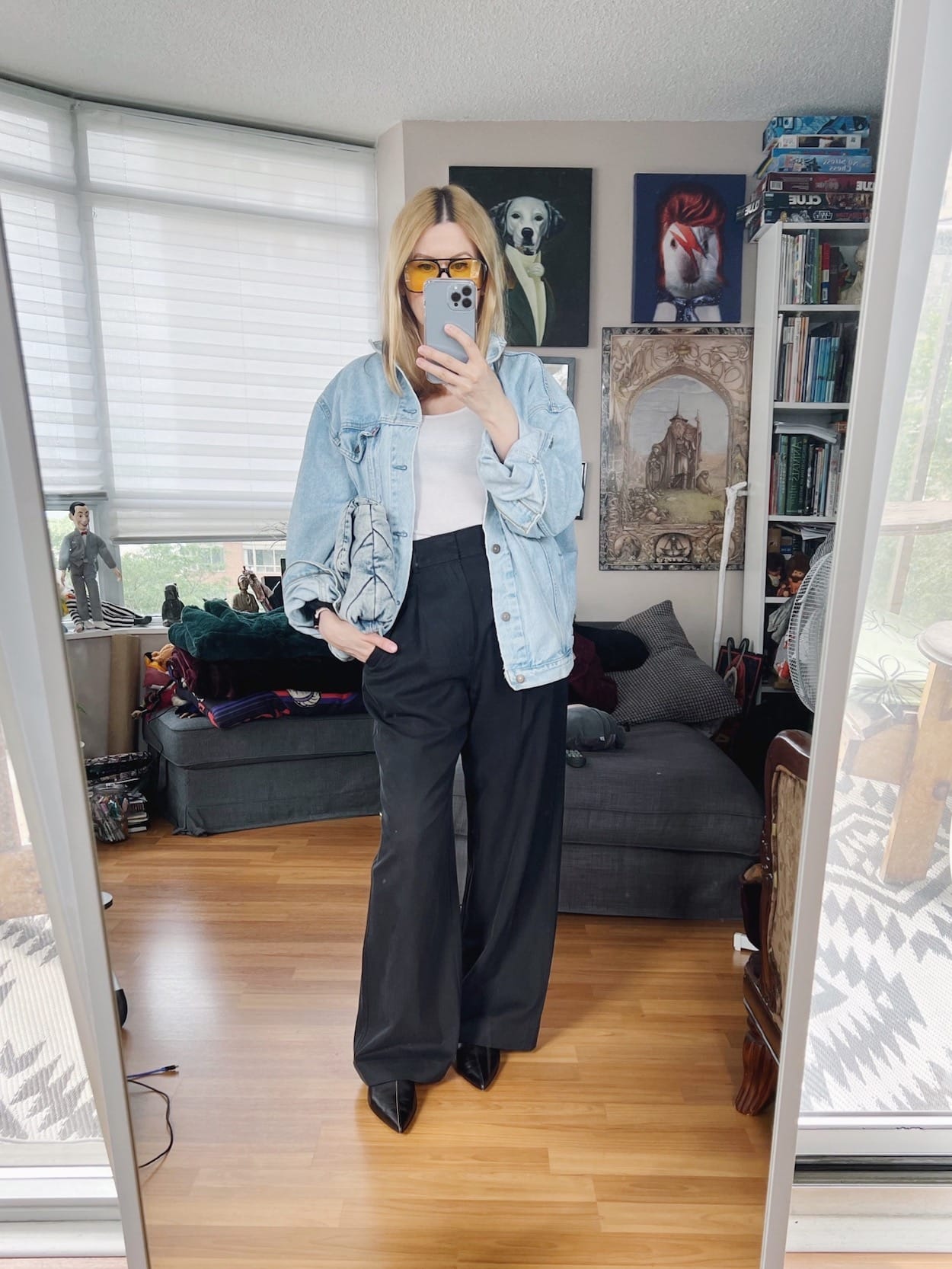 A blonde woman is wearing a white tank, vintage jean jacket, trousers, flats, a vintage bag, and retro sunglasses.