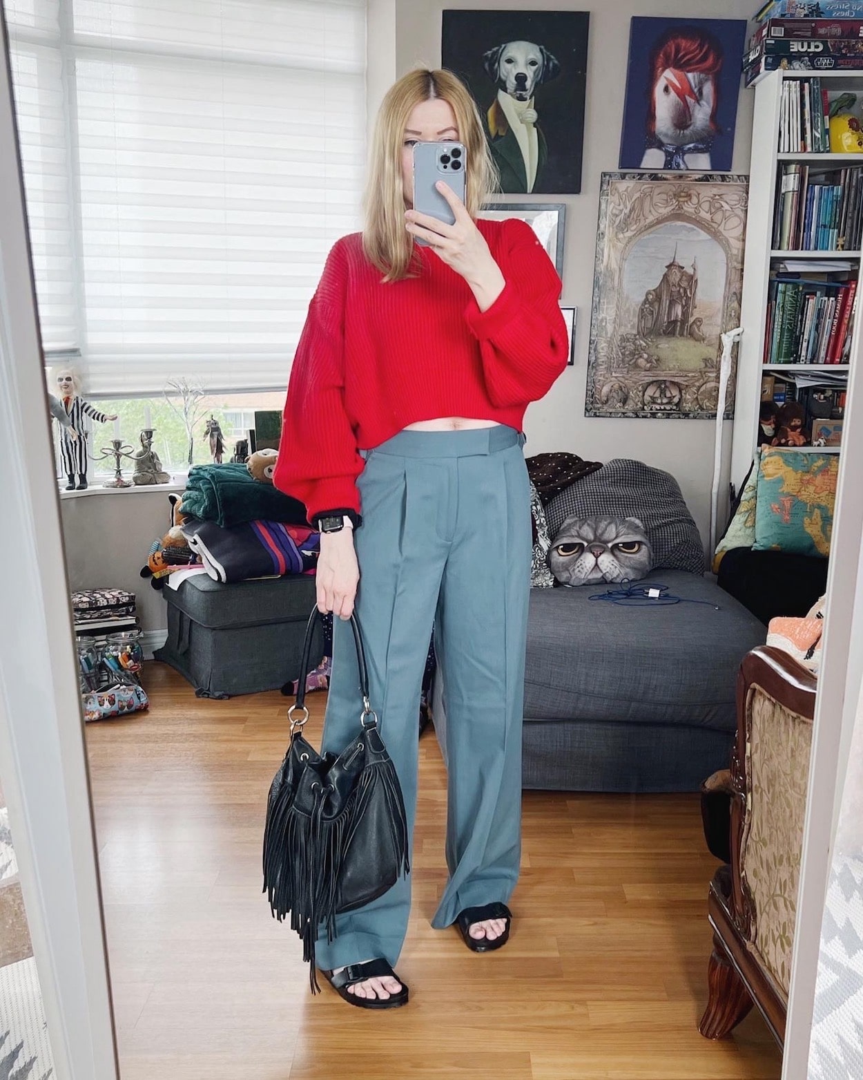 A blonde woman is wearing a red cropped sweater, blue trousers, Birkenstocks, and a vintage Roots bucket bag.