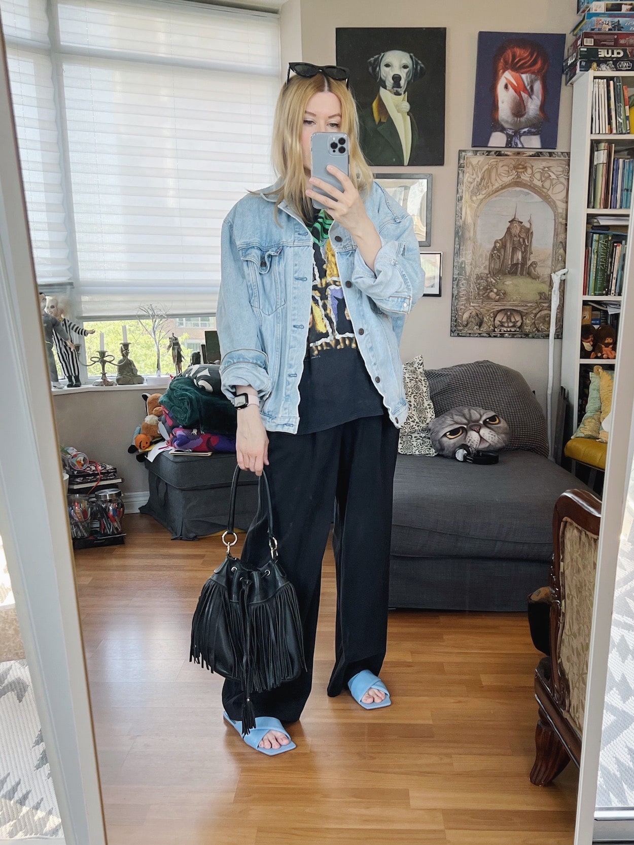 A blonde woman is wearing a vintage Poison t-shirt, vintage Levi's jacket, black trousers, blue sandals, sunglasses, and a vintage bucket bag.