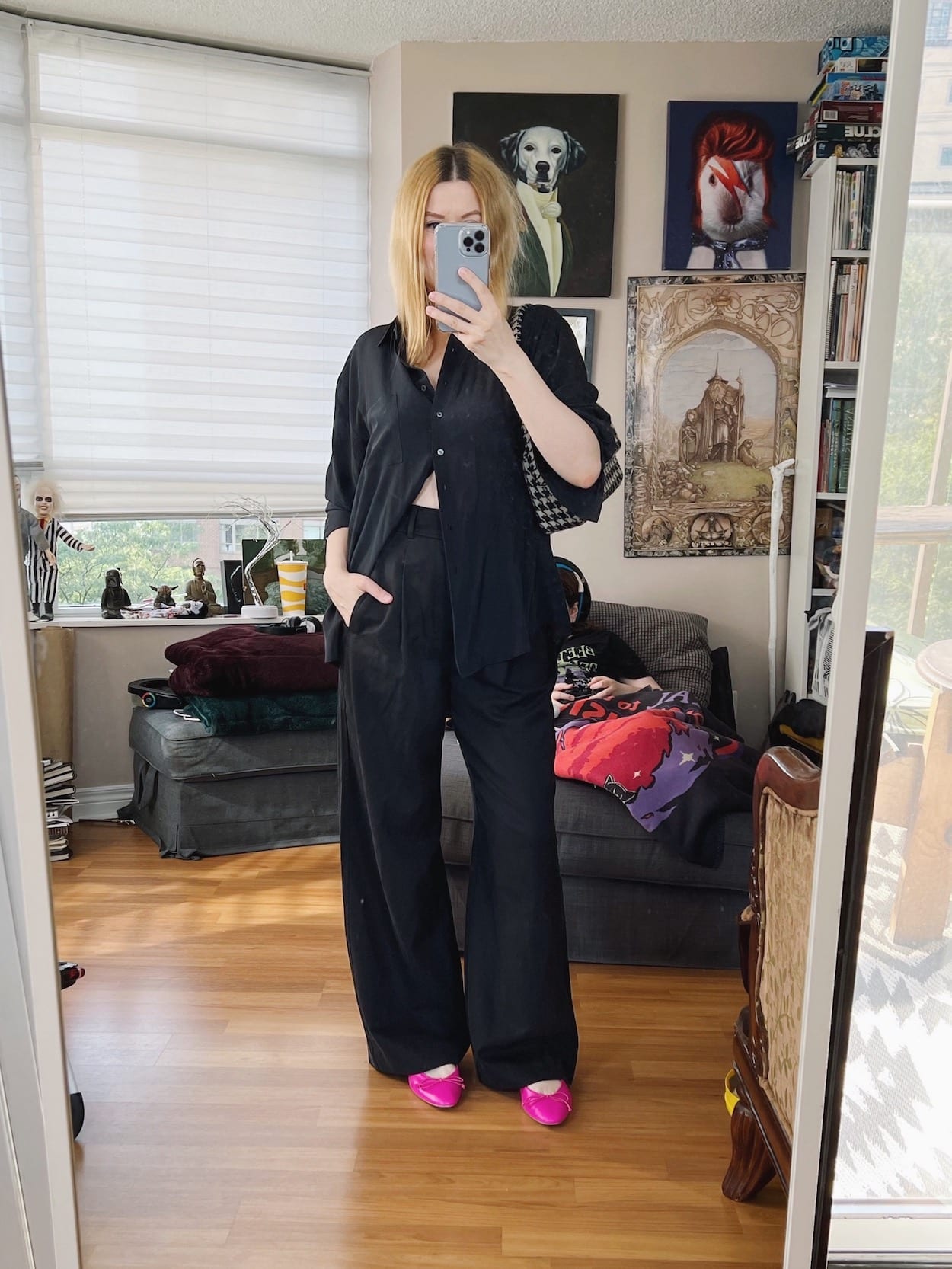 A blonde woman is wearing a black silk blouse, black trousers, pink ballet flats, and an Anine Bing bag.