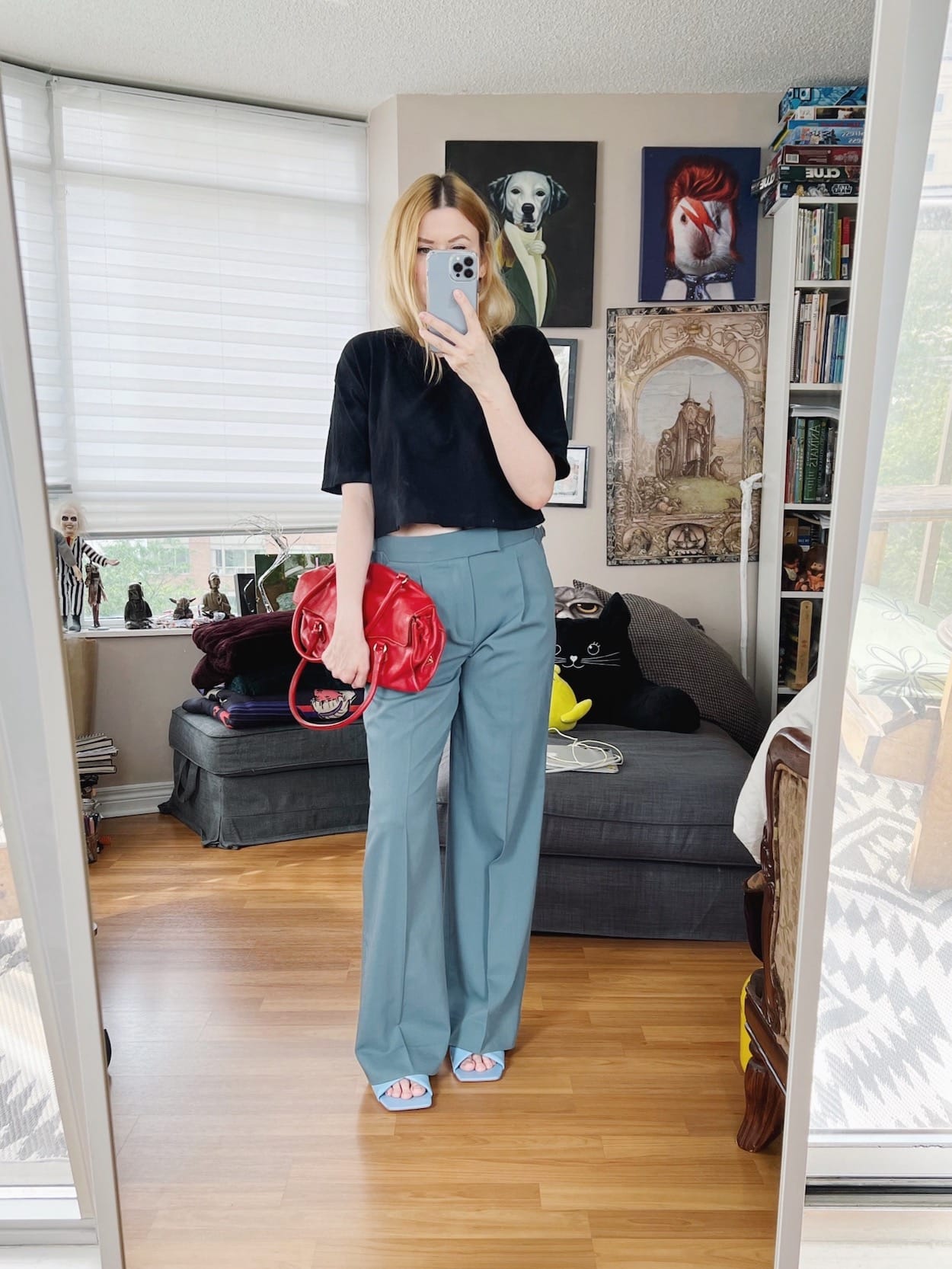 A blonde woman is wearing a black cropped t-shirt, blue trousers, blue sandals, and a red vintage purse.