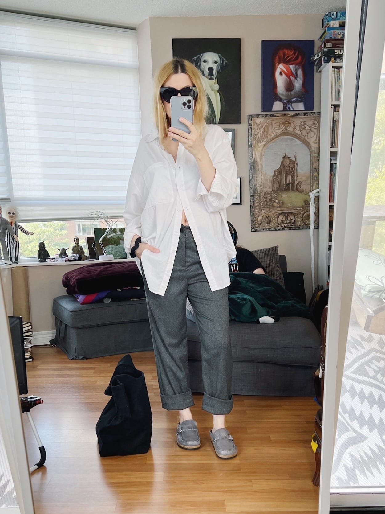 A blonde woman is wearing a white button up, grey trousers, Birkenstock clogs, LeSpec sunglasses, and a black tote.
