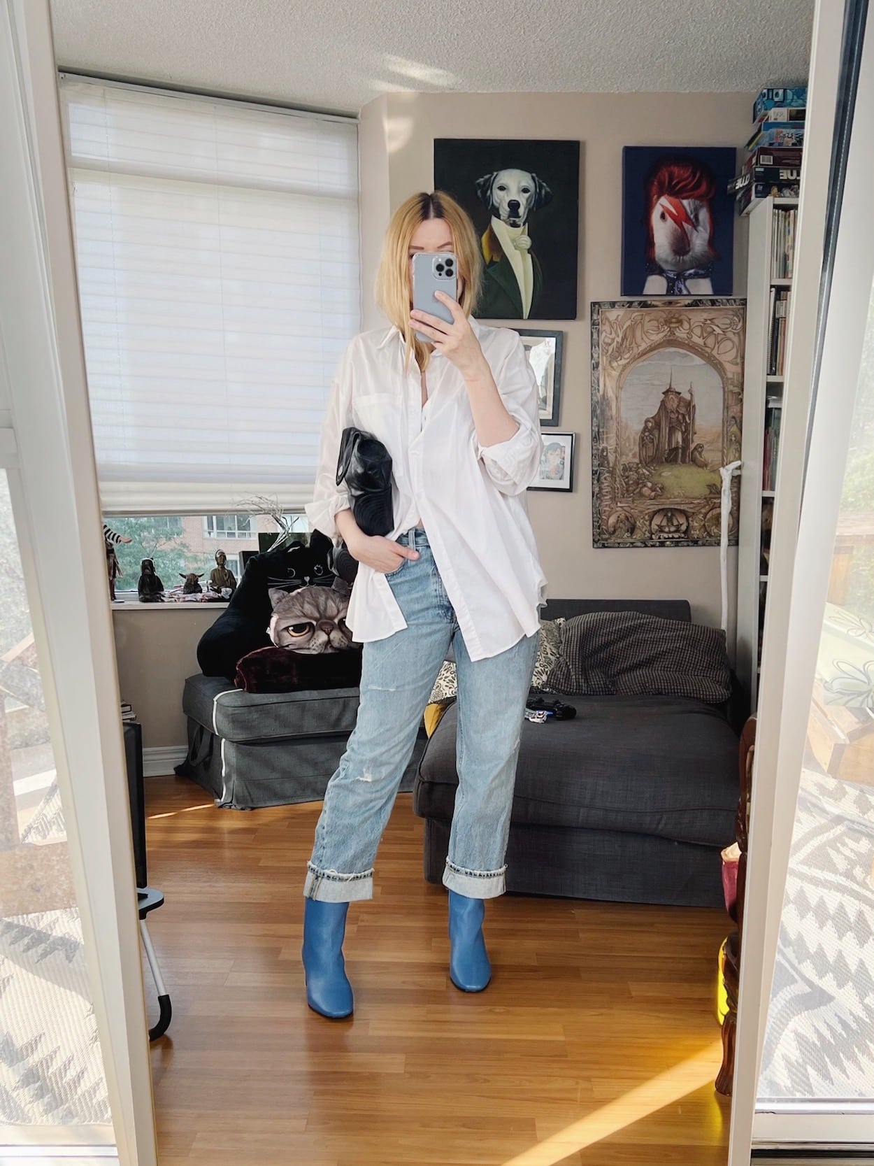 A blonde woman is wearing a white button up, jeans, blue boots, and is carrying a large clutch.