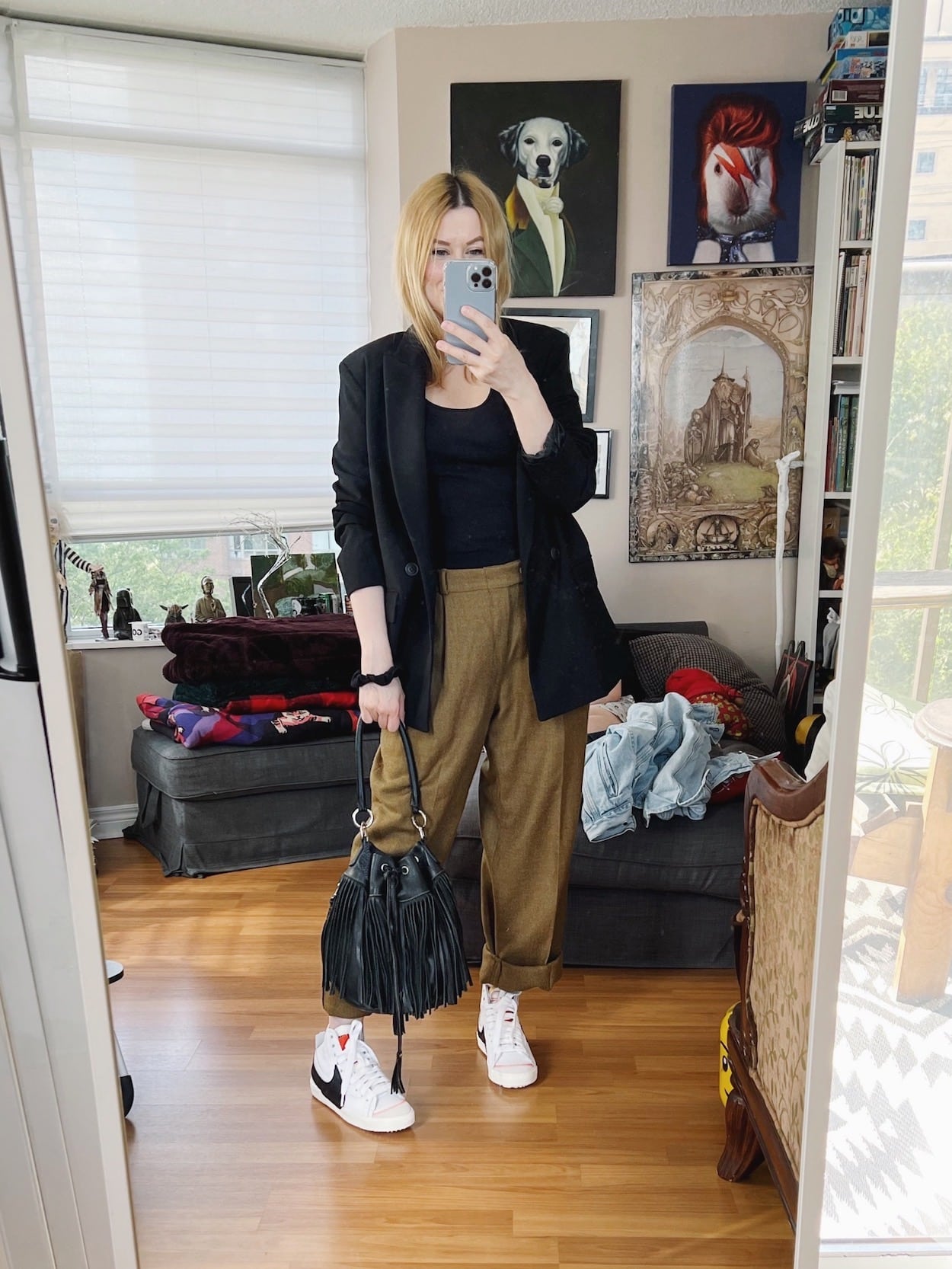A blonde woman is wearing vintage trousers, a black tank, black blazer, Nikes, and a vintage fringe bucket bag.