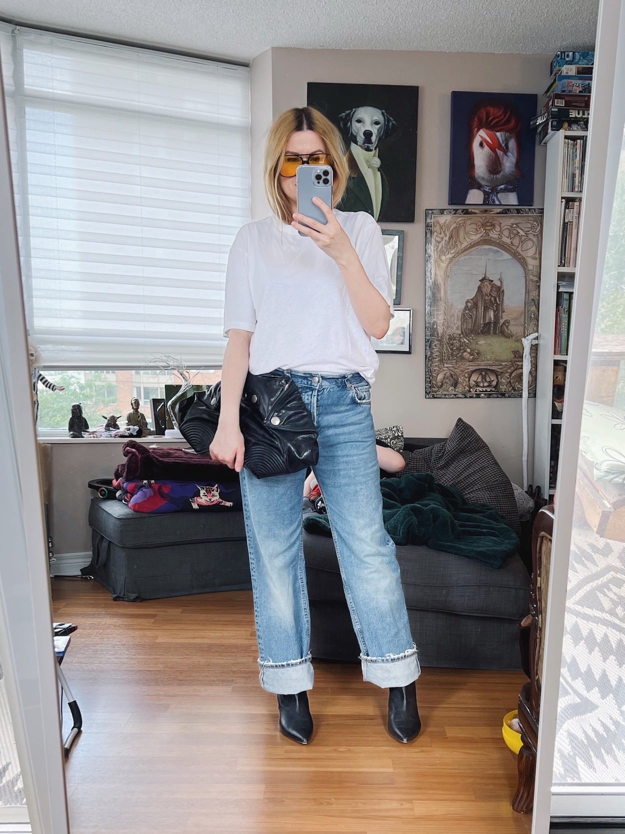 A blonde woman is wearing a white t-shirt, zara boyfriend jeans, knee high boots (under jeans), retro sunglasses, and an Alexander McQueen clutch.