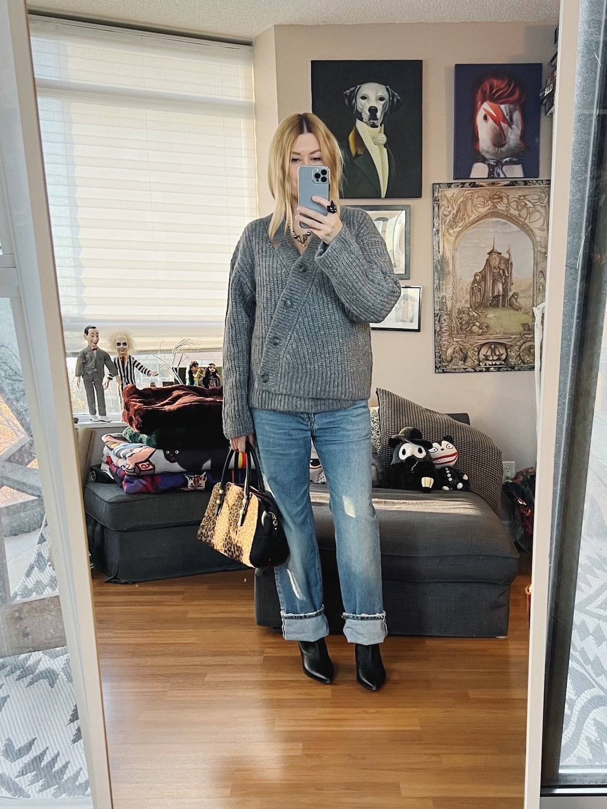 A blonde woman is wearing a grey cardigan sweater, boyfriend jeans, black boots, and is carrying an animal print bag.