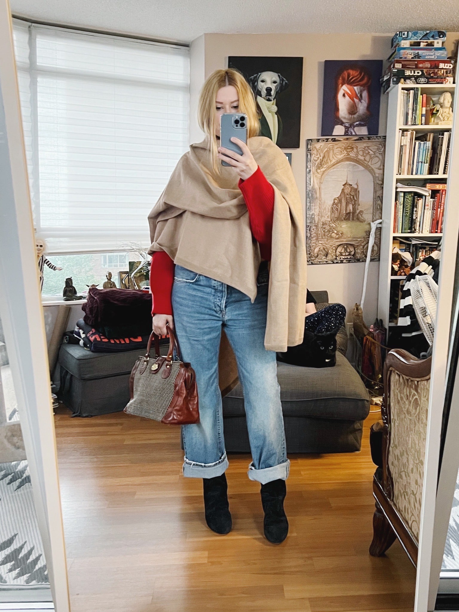 A blonde woman is wearing a red sweater, a cape, boyfriend jeans, boots, and a vintage bag.