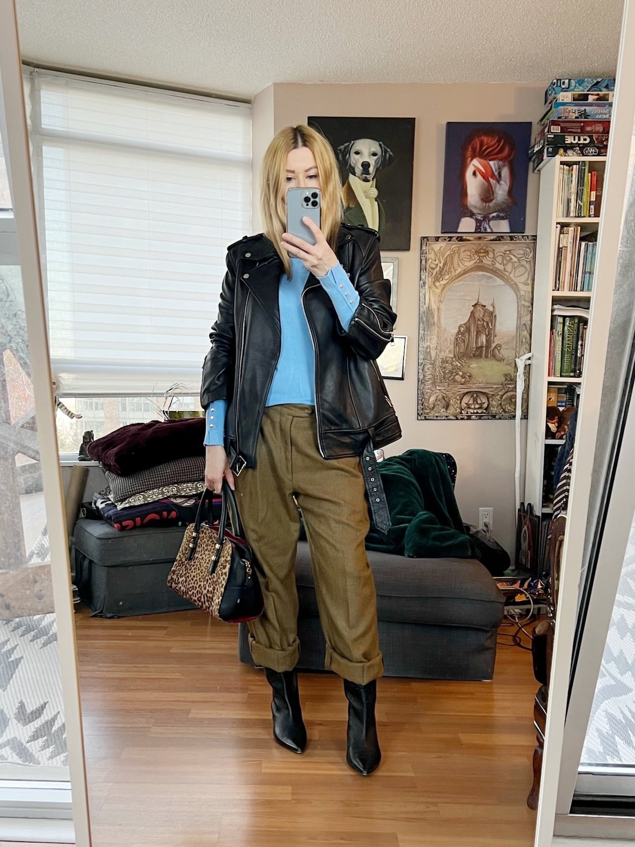 A blonde woman is wearing a light blue sweater, brown vintage trousers, knee high boots, a leather jacket, and is carrying an animal print bag.