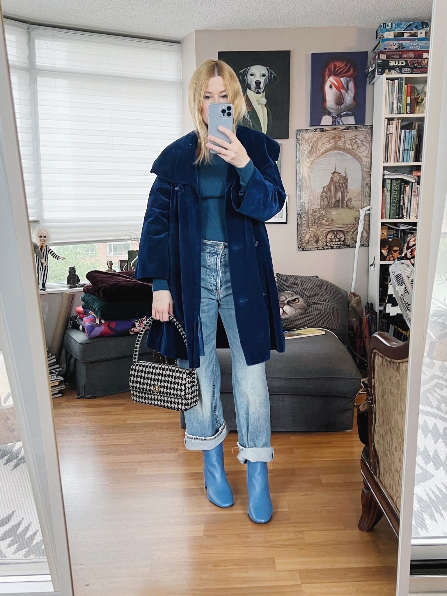 A blonde woman is wearing a blue turtleneck, boyfriend jeans, a blue velvet coat, blue boots, and a houndstooth bag.