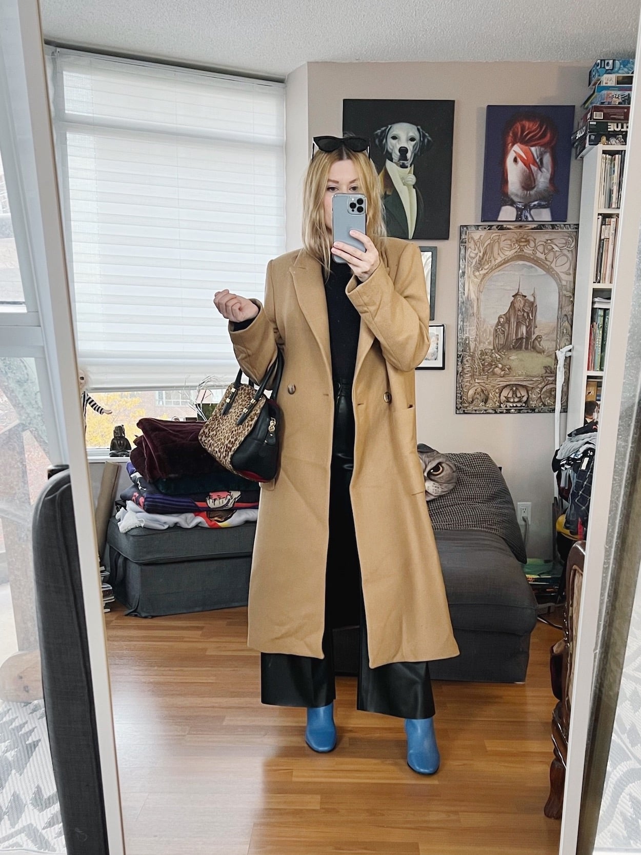 A blonde woman is wearing a black turtleneck, faux leather pants, a long camel coat, blue boots, sunglasses, and is carrying an animal print bag.