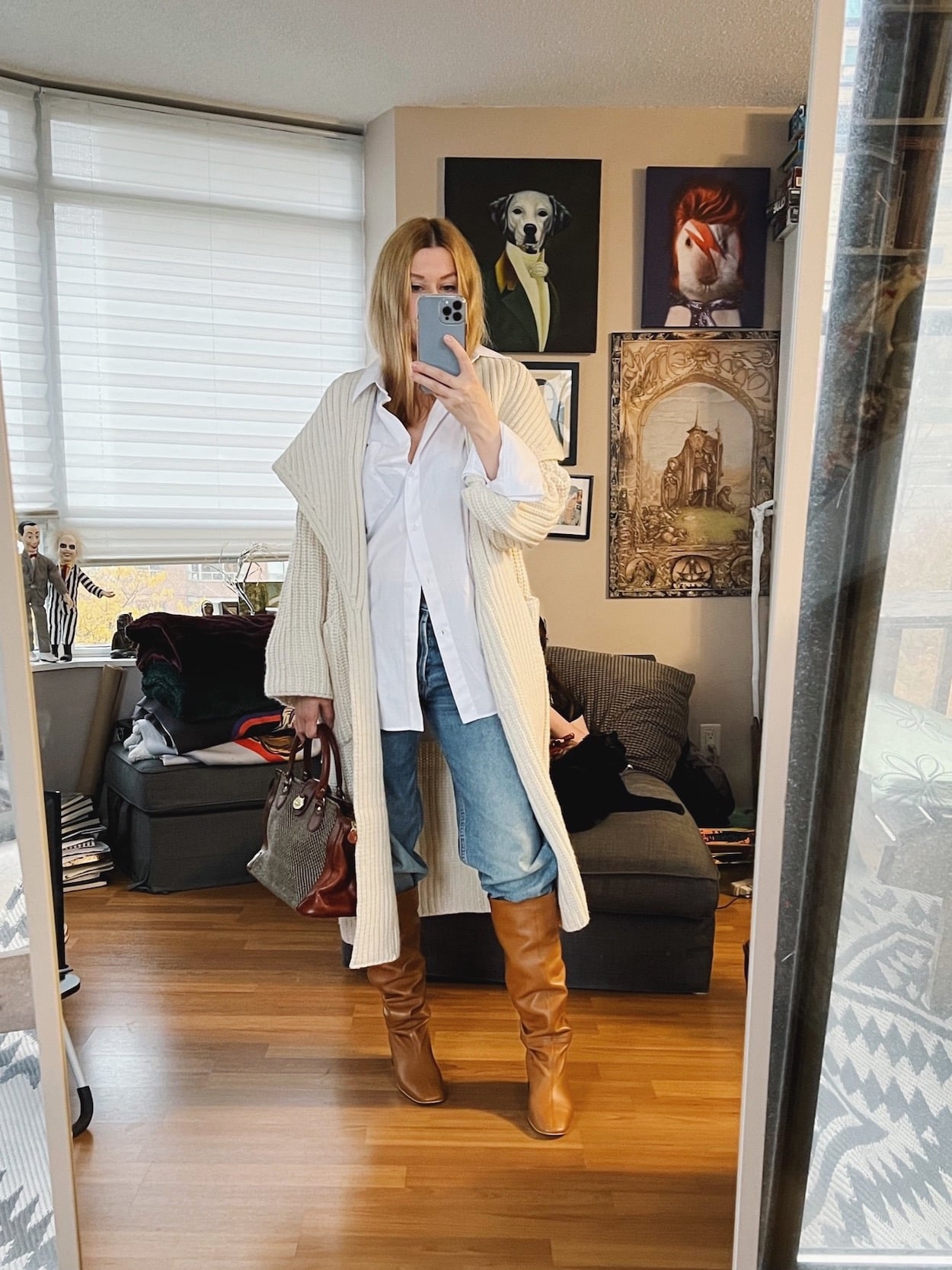 A blonde woman is wearing a white button up with a long cream cardigan, boyfriend jeans, brown knee high boots, and a vintage bag.