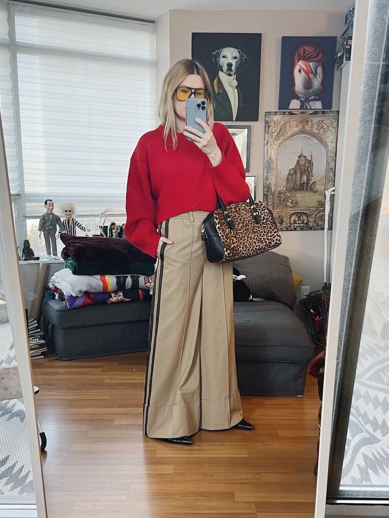 A blonde woman is wearing a cropped red sweater, wide leg trousers, pointy toe flats, retro sunglasses, and an animal print bag.