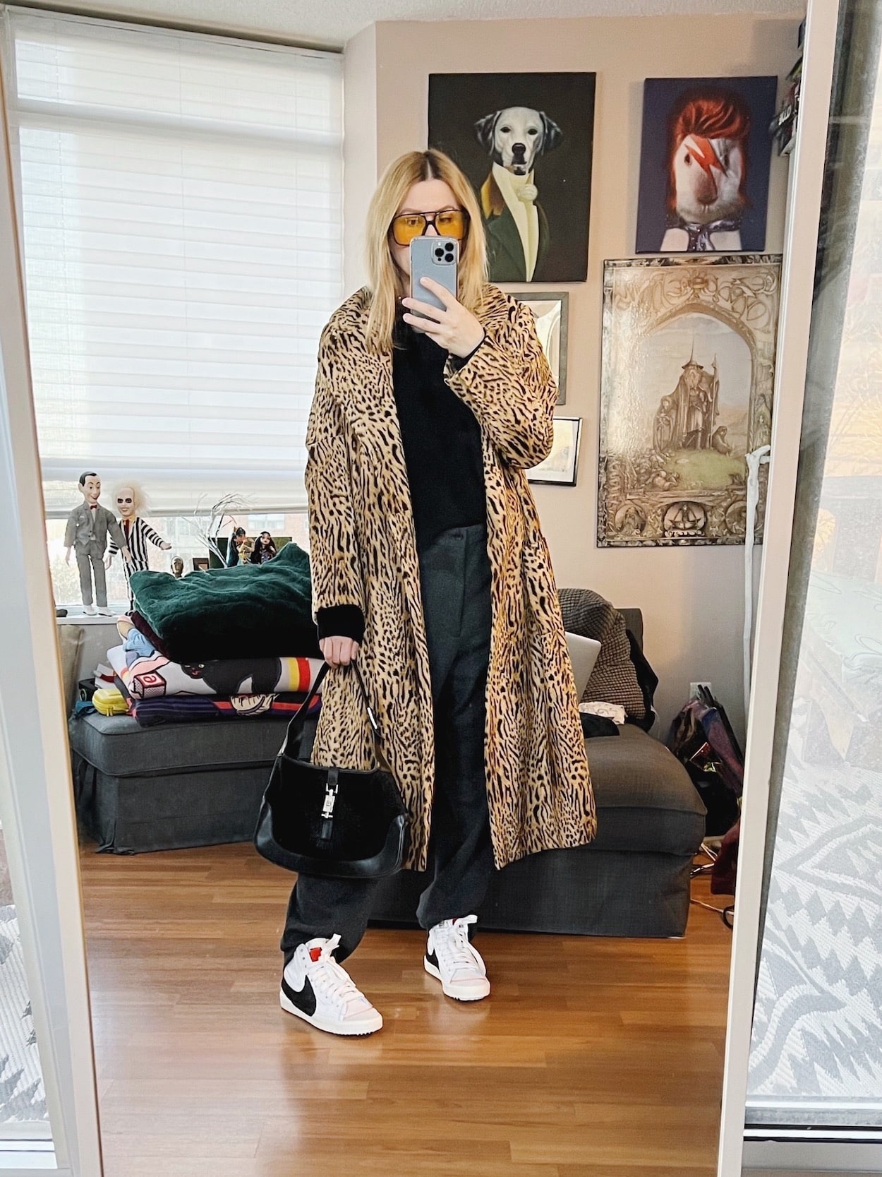 A blonde woman is wearing a vintage animal print coat, grey trousers, a black sweater, Nikes, retro sunglasses, and a vintage Gucci bag.