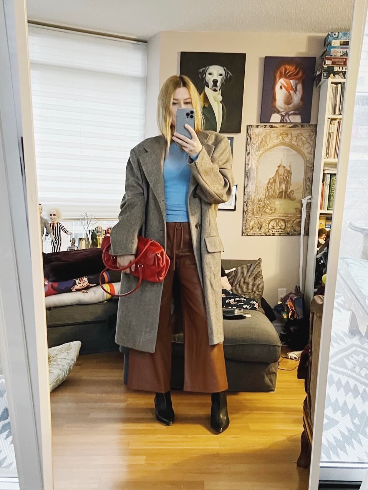 A blonde woman is wearing a blue sweater, brown leather pants, black boots, an oversized men's coat, and a red vintage Prada bag.