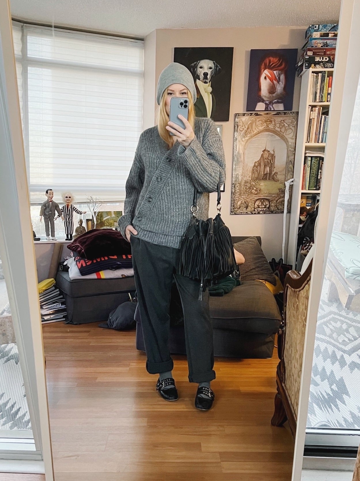 A blonde woman is wearing a grey sweater, men's trousers, ballet flats, a grey beanie, and a vintage bucket bag.