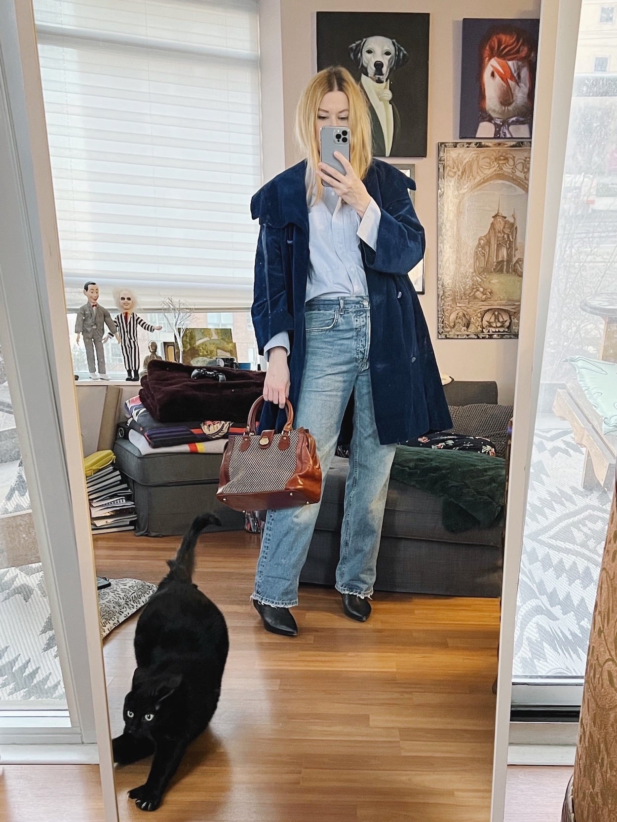 A blonde woman is wearing a blue button-up, jeans, black boots, vintage blue velvet coat, and a vintage bag.