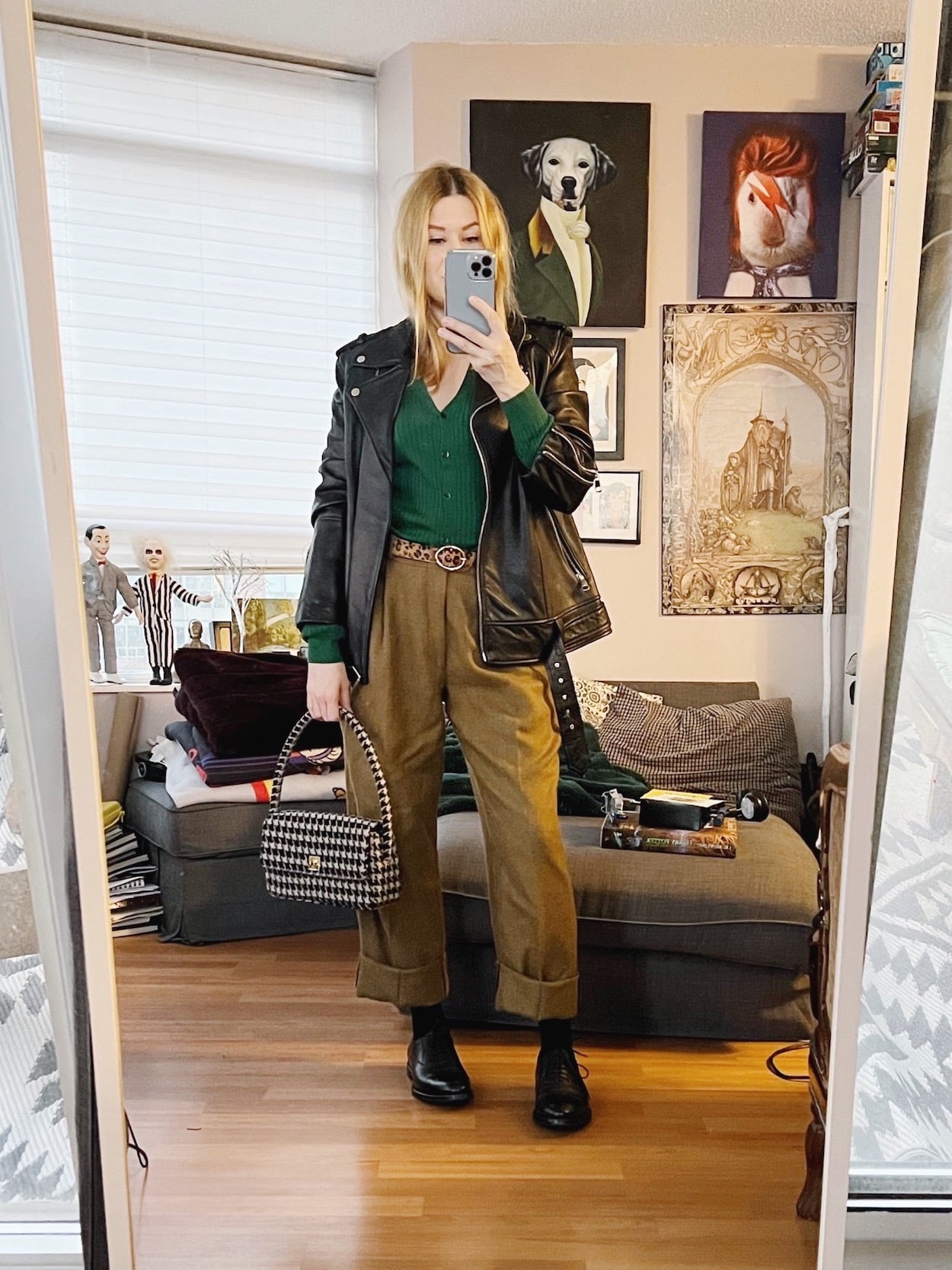 A blonde woman is wearing a green cardigan, men's trouser, an animal print belt, oversized leather jacket, derby shoes, and a houndstooth bag.