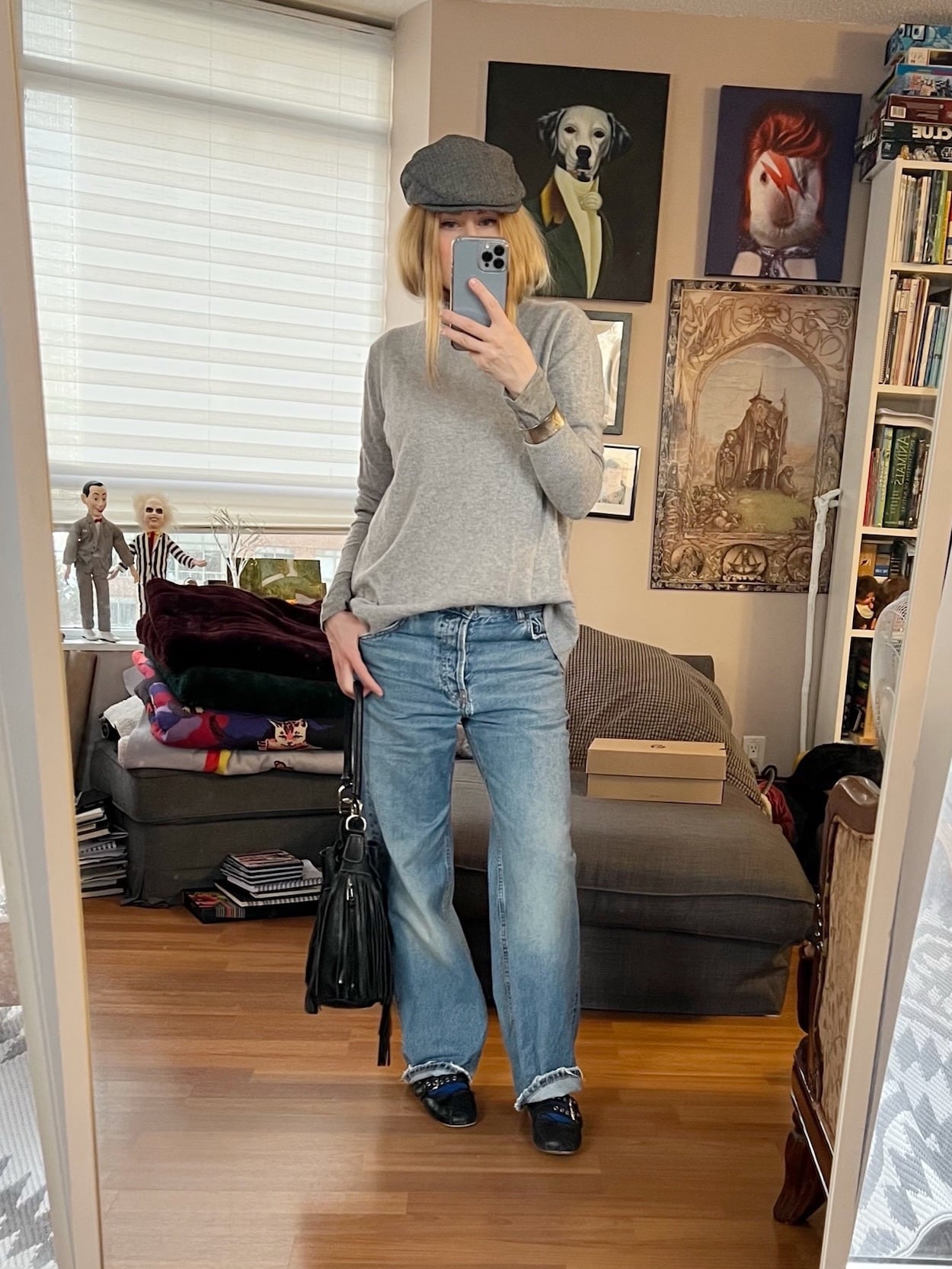 A blonde woman is wearing a grey cashmere sweater. boyfriend jeans, Miu Miu flats, a driving cap, and a vintage bucket bag.