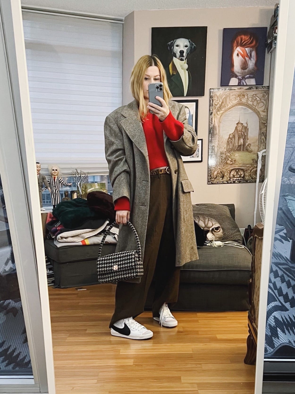 A blonde woman is wearing a red cropped sweater, men's vintage trousers, a vintage men's overcoat, Nike Blazers, and a houndstooth bag.