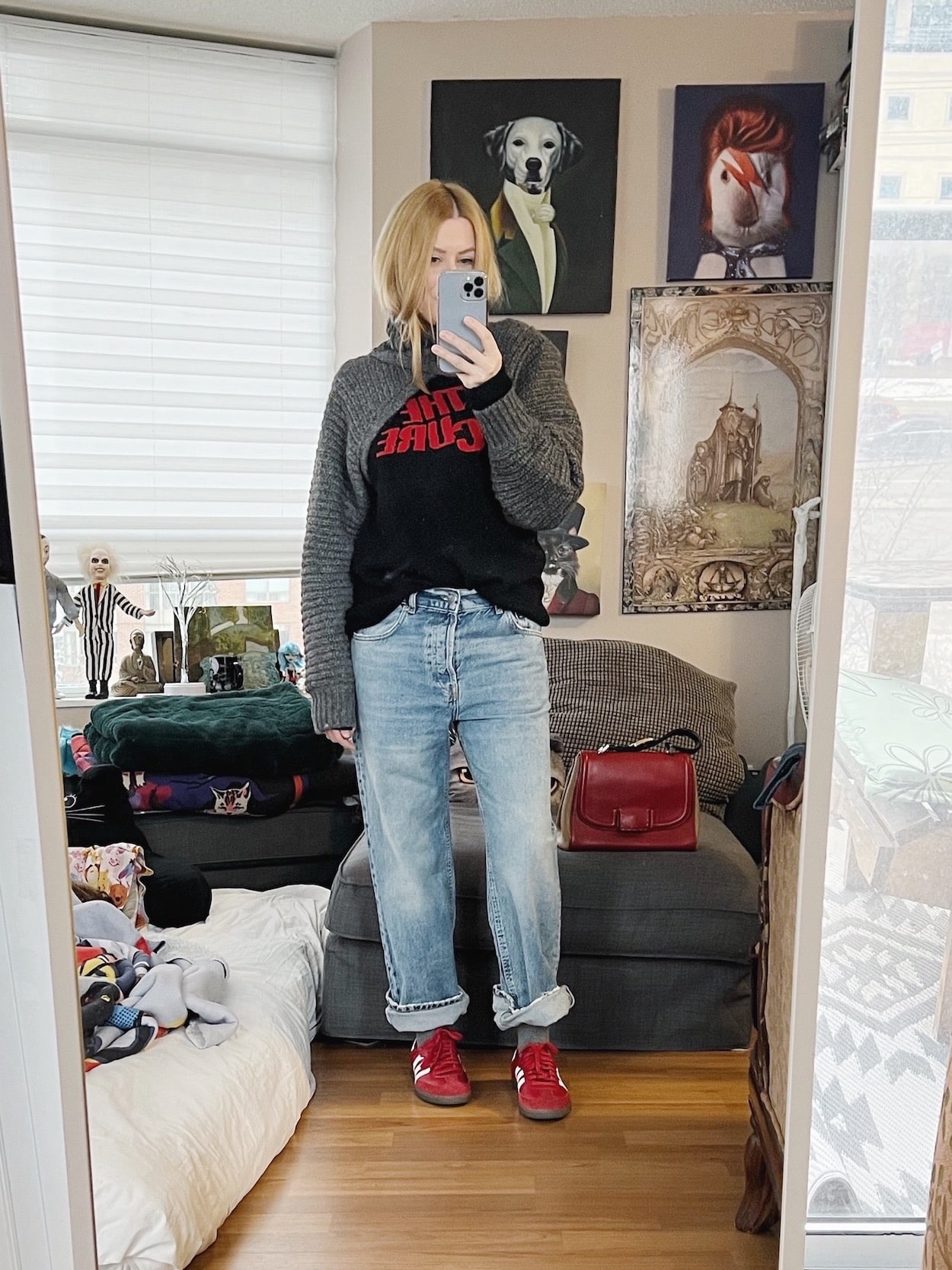A blonde woman is wearing A Cure sweater, shrug, jeans, and red sneakers.