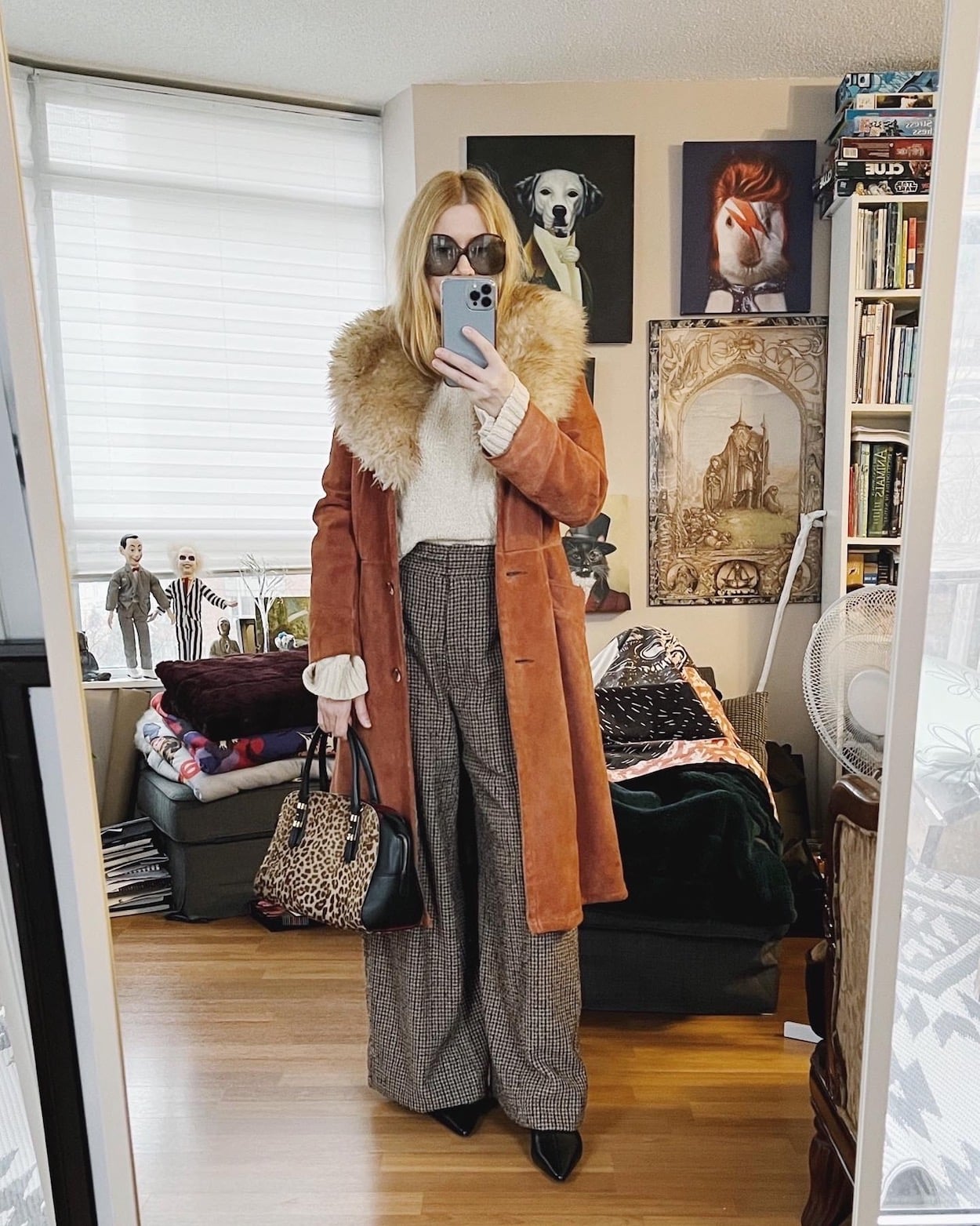 A blonde woman is wearing wide leg trousers, a sweater, a vintage coat, kitten heel boots, vintage sunglasses, and an animal print bag.