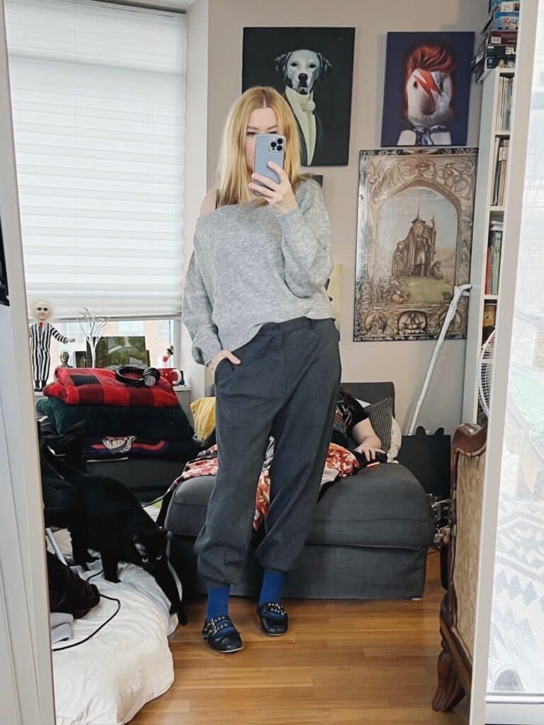 A blonde woman is wearing a grey off the shoulder sweater with slouchy trousers, and ballet flats.