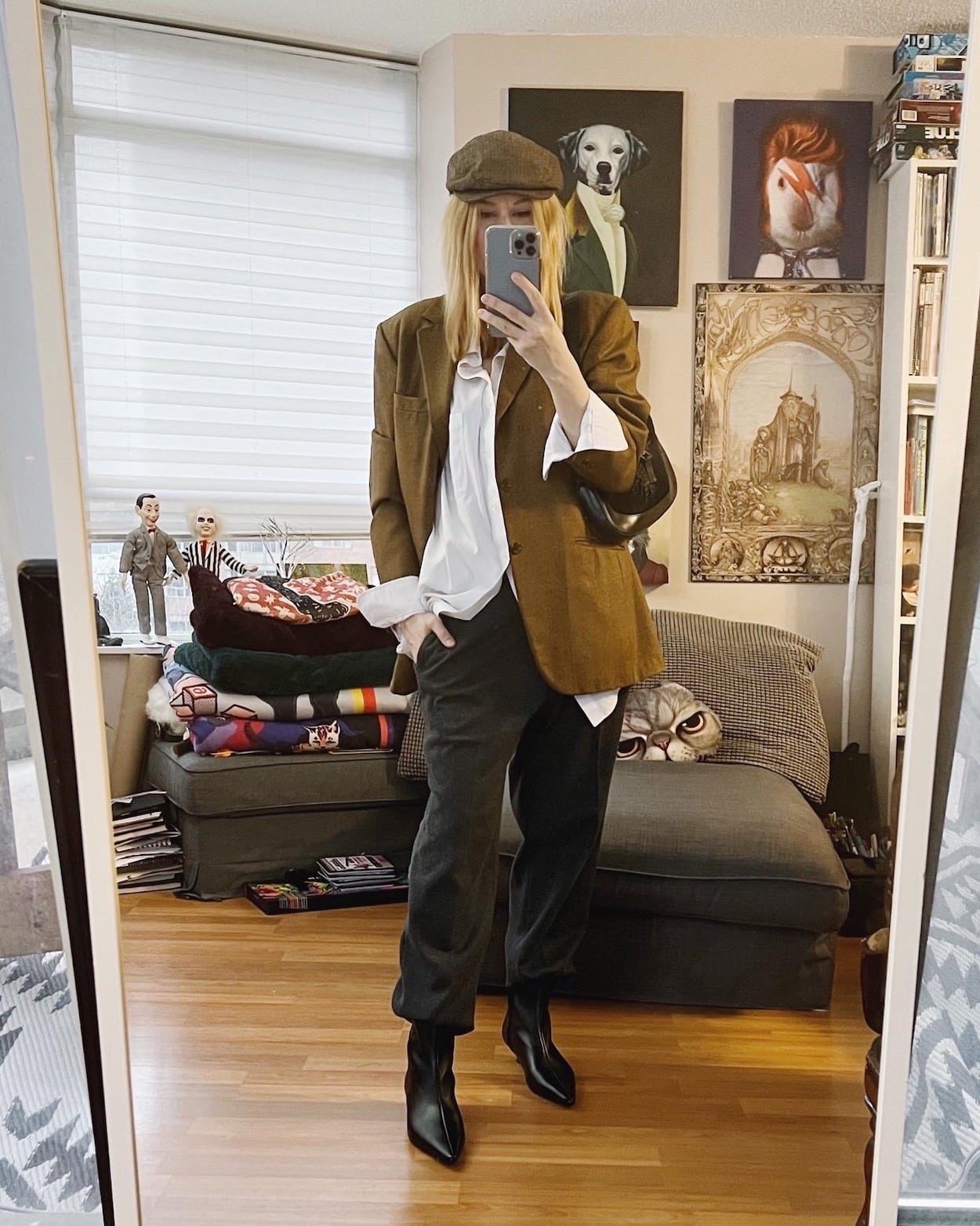 A blonde is wearing a button up, oversized blazer, men's trousers, kitten heel boots, and a driving hat.