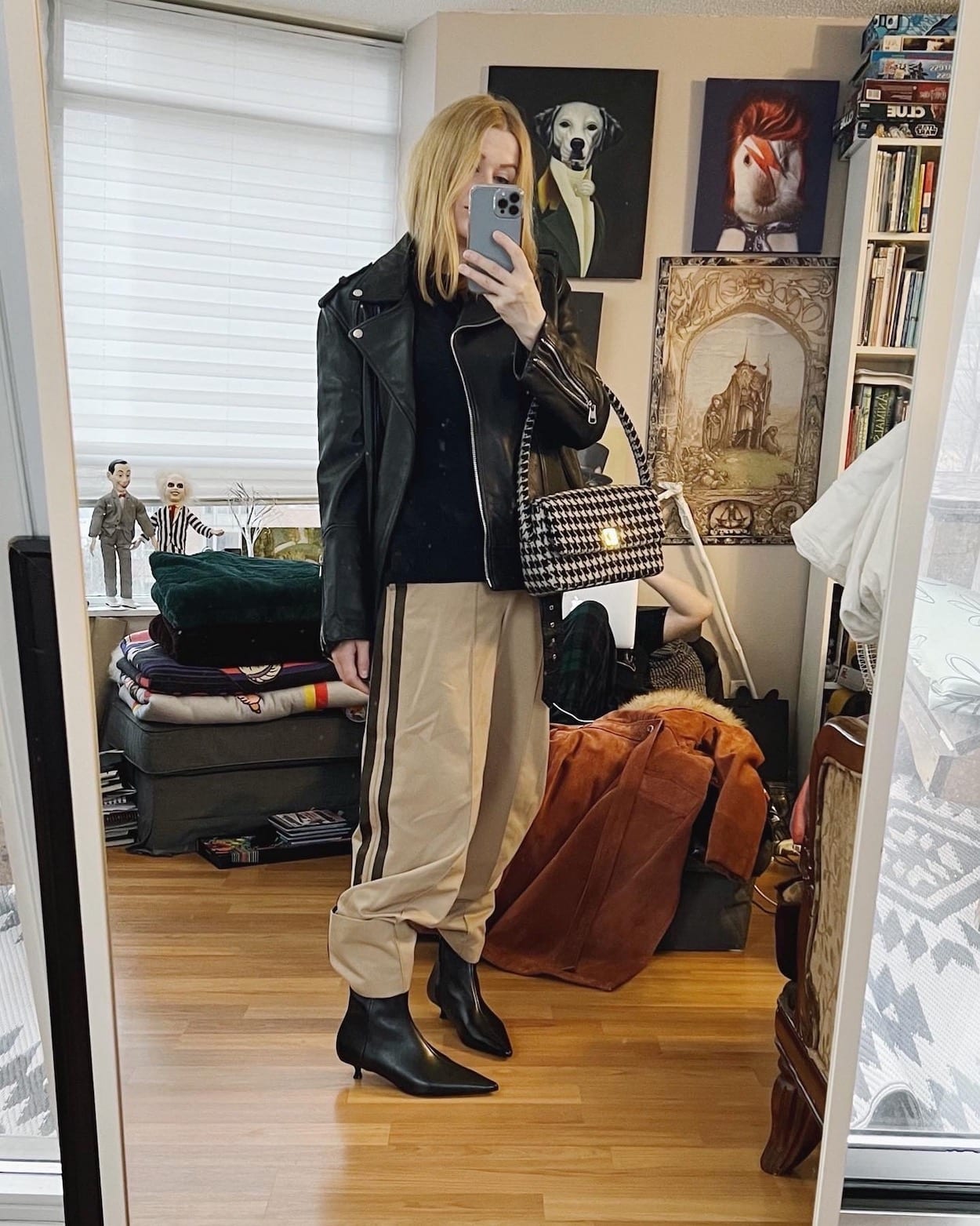 A blonde woman is wearing trousers, kitten heel boots, a black sweater, oversized leather jacket, and a houndstooth bag.