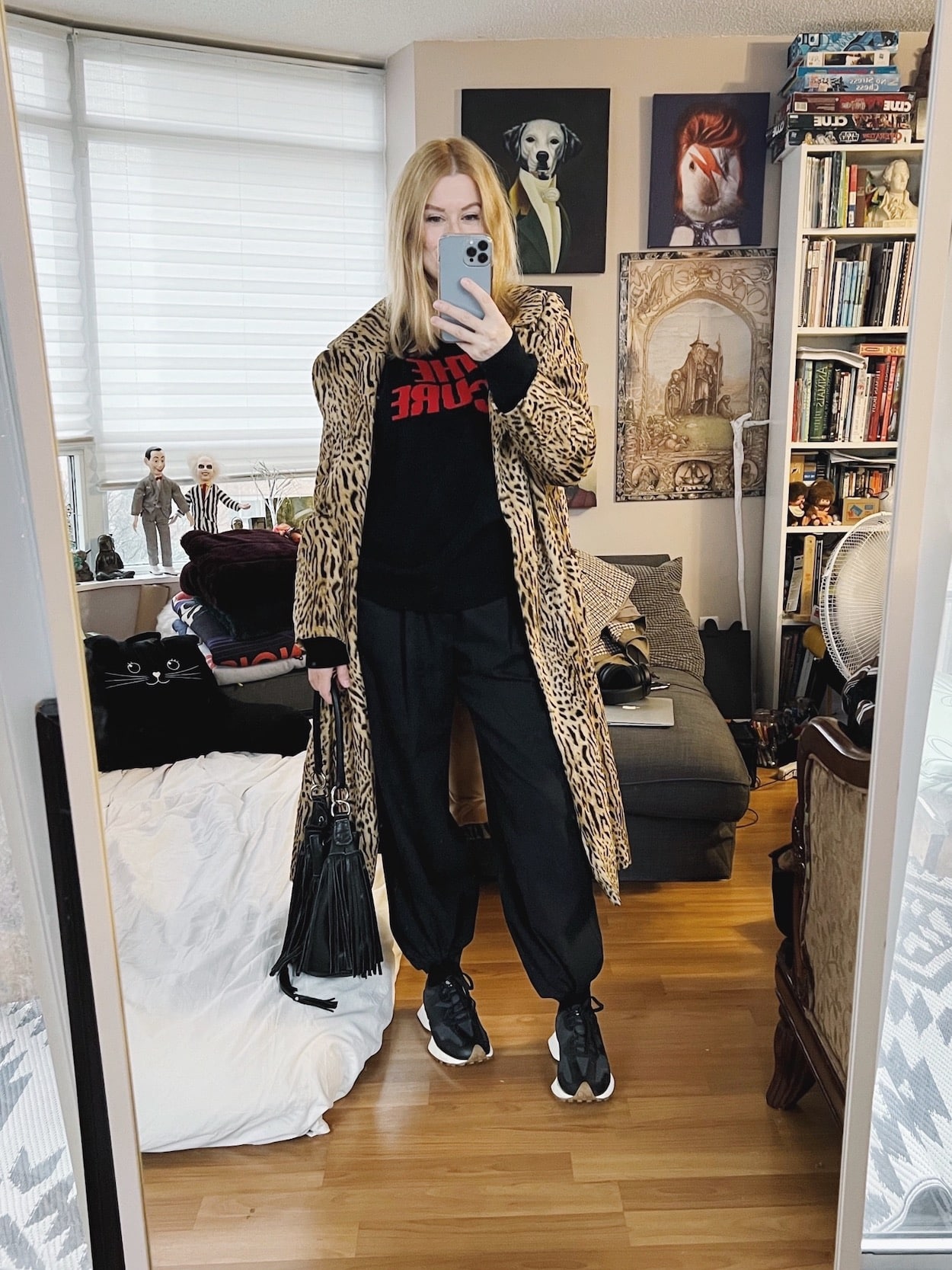 A blonde woman is wearing black trosuers, The Cure sweater, a vintage animal print coat, sneakers, and a fringe bucket bag.