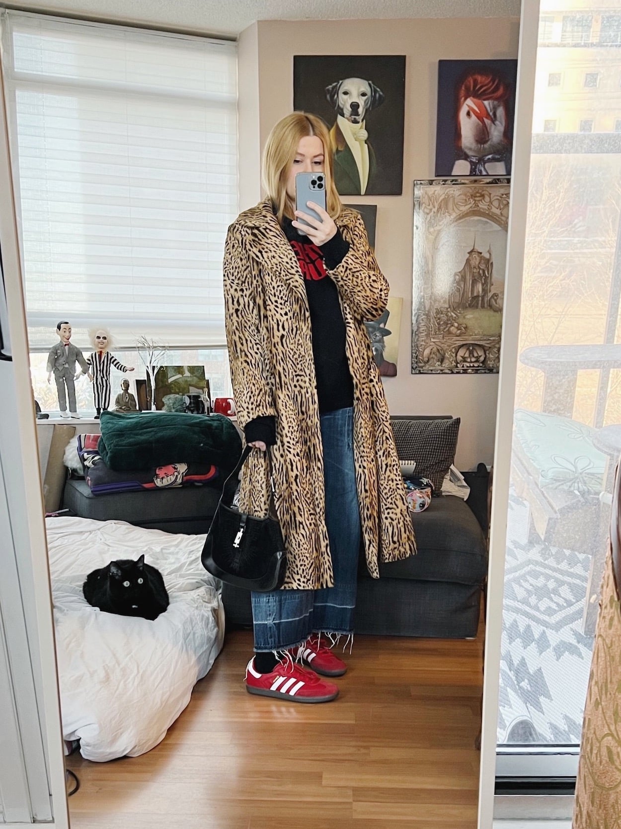 A blonde woman is wearing a Cure sweater, vintage coat, wide leg jeans, a vintage bag and red Sambas.
