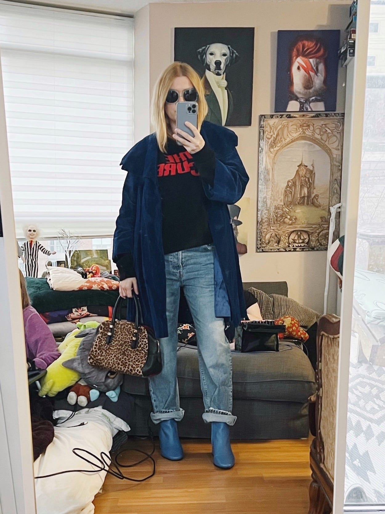 A blonde woman is wearing a knit band sweater, boyfriend jeans, a vintage swing coat, blue boots, an animal print bag, and vintage sunglasses.